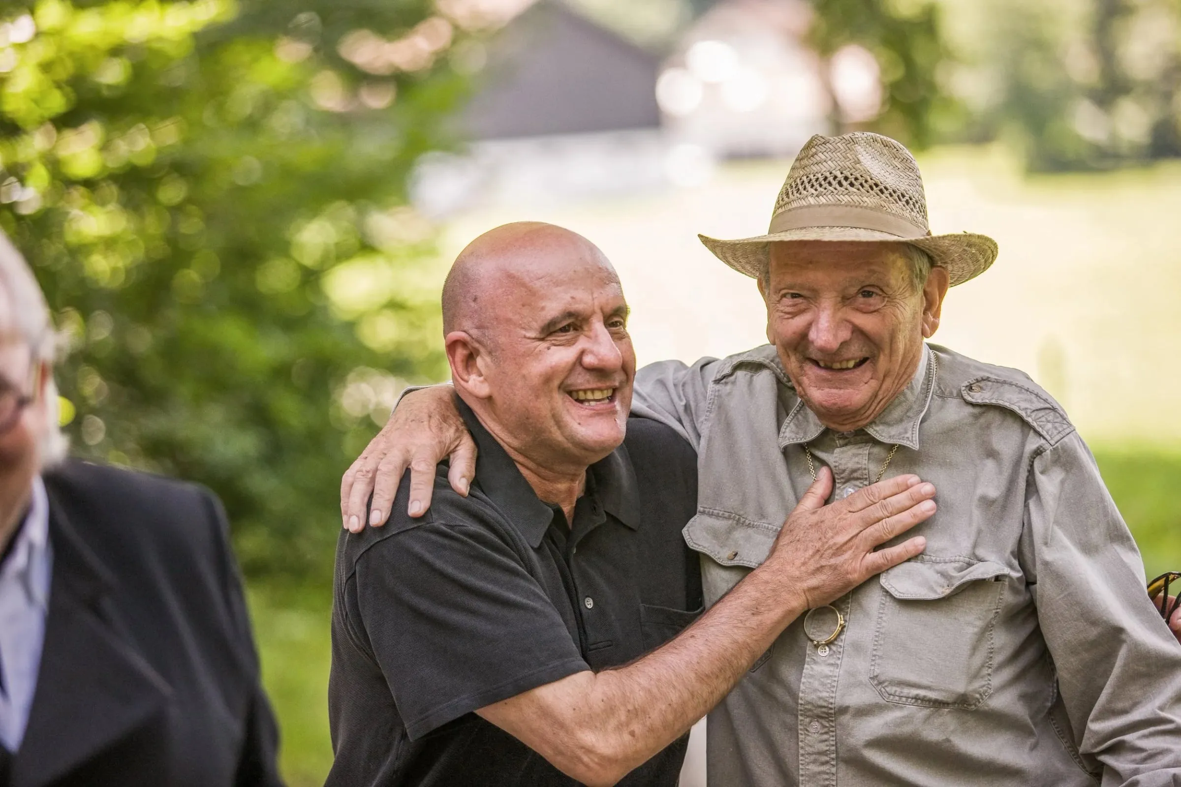 Zwei Männer im Freien Arm in Arm, beide lachen in die Kamera, grüner Hintergrund