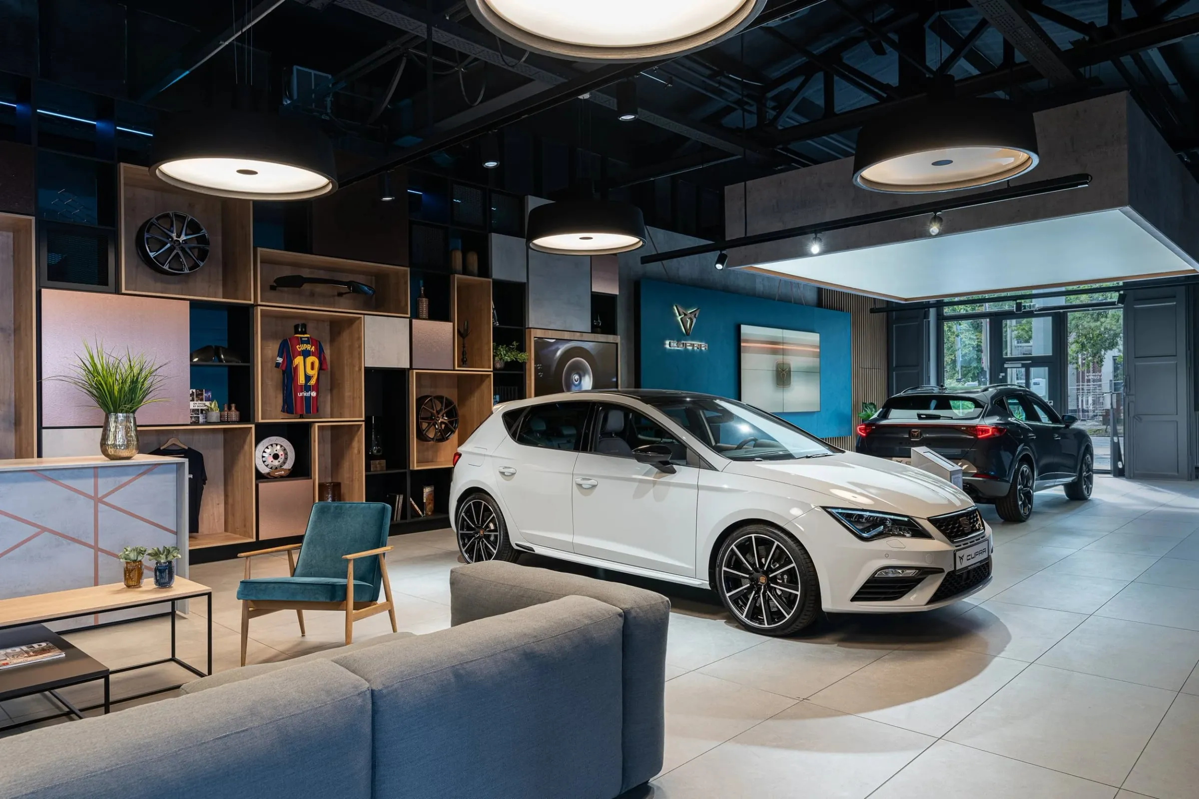 Weißer Cupra mit dunklen Felgen mittig im Showroom, runde Deckenleuchten, Holzregale und Sitzbereich seitlich