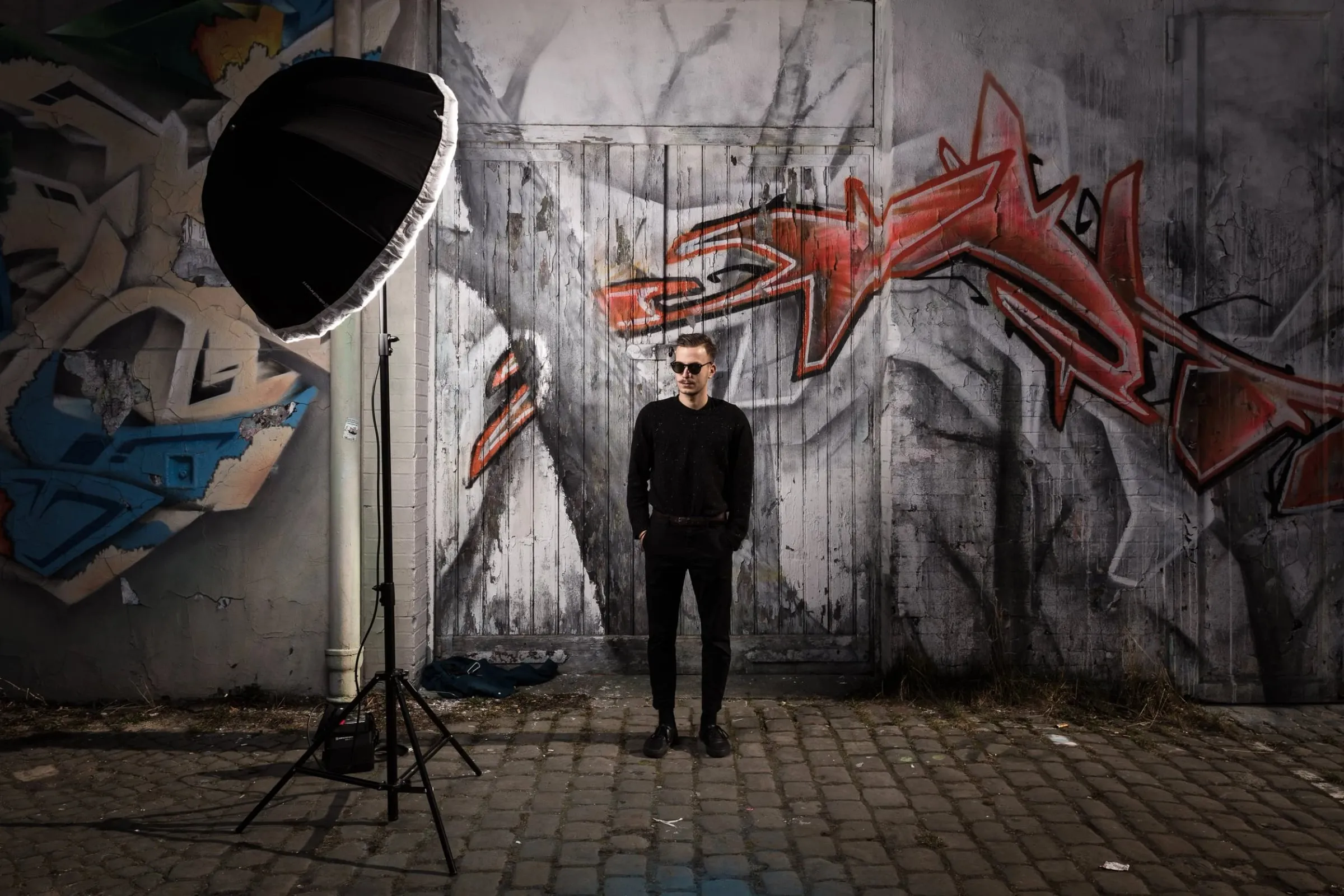 Junger Mann in Schwarz mit Sonnenbrille vor einer Graffitiwand, Studioblitz mit Softbox links, Ganzkörperaufnahme draußen
