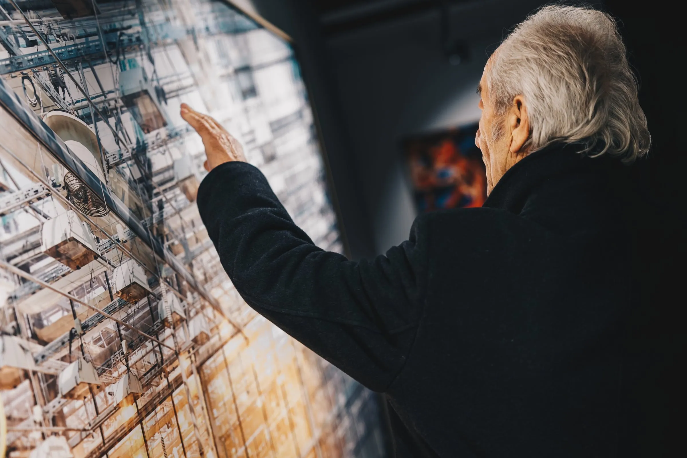Älterer Mann deutet mit der Hand auf eine großformatige gerahmte Architekturfotografie an der Wand