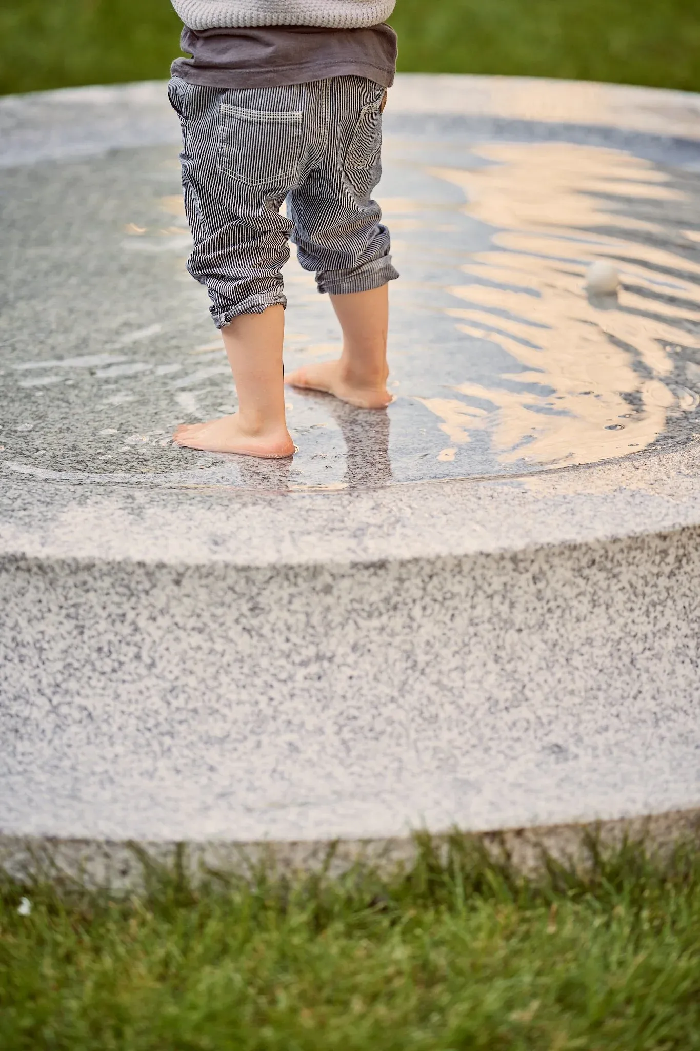 Barfüßiges Kind steht auf dem flachen Wasserrand eines runden Granitbrunnens, umgeben von Rasen