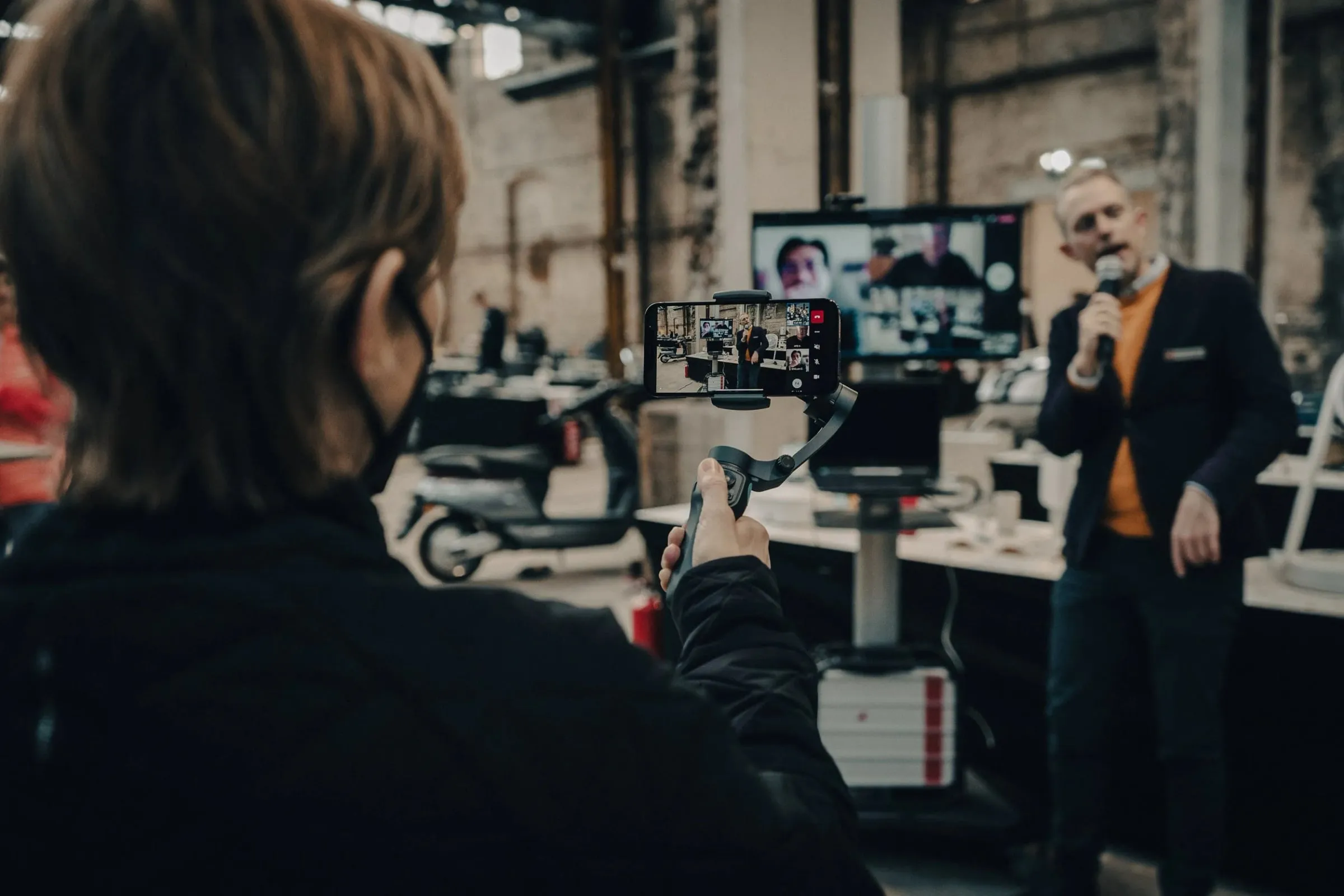 Mann mit Mikrofon spricht auf der Bühne, im Vordergrund filmt eine Person mit Smartphone am Gimbal