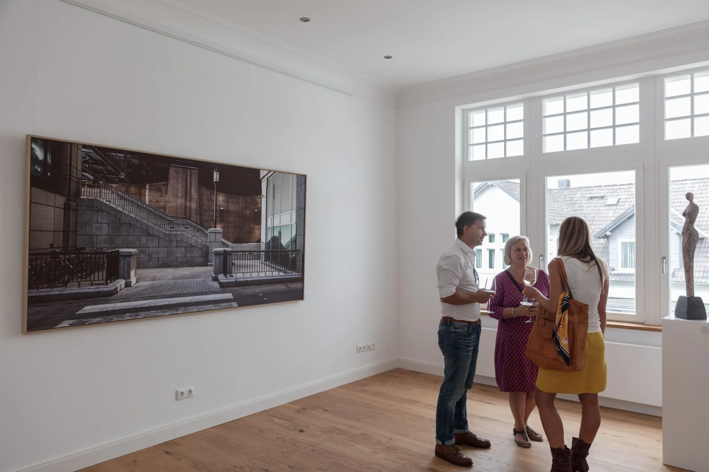 Drei Gäste vor großformatiger gerahmter Fotografie einer Treppe, daneben Sprossenfenster und Skulptur