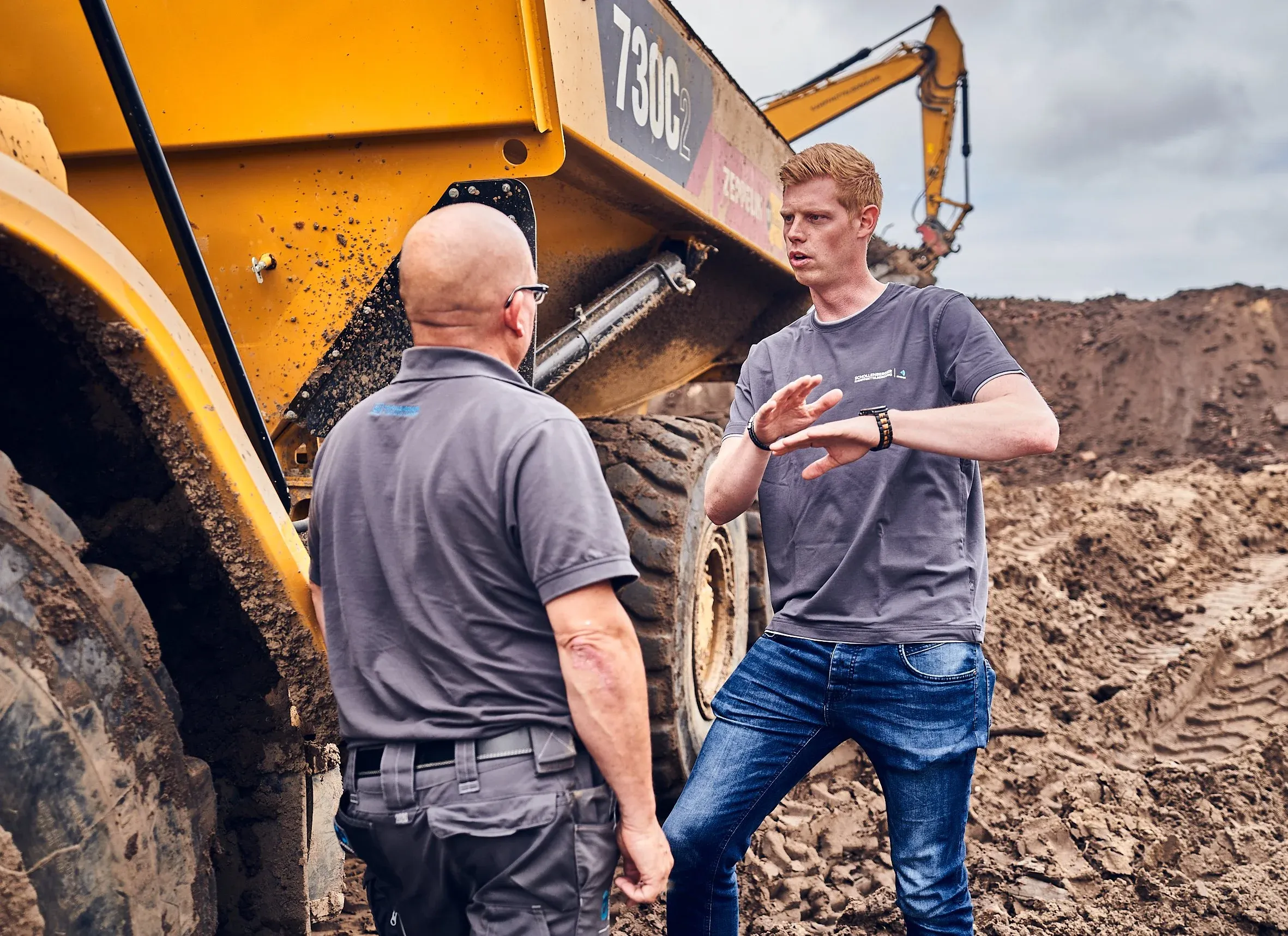 Zwei Arbeiter im Gespräch vor gelbem Muldenkipper, im Hintergrund ein Bagger auf Sand