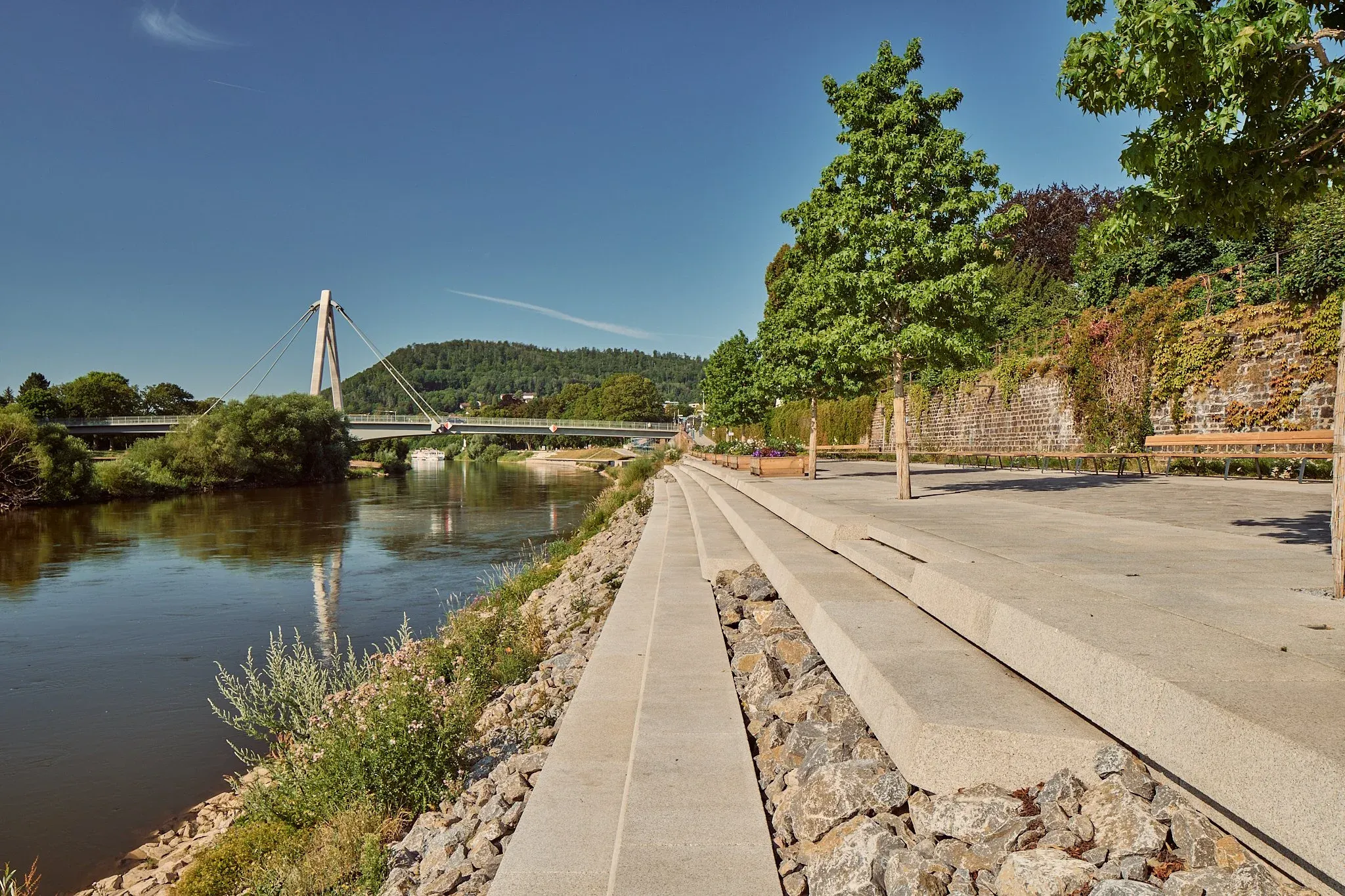 Uferweg an der Weser bei Sonnenschein, Schrägseilbrücke und bewaldete Hügel im Hintergrund