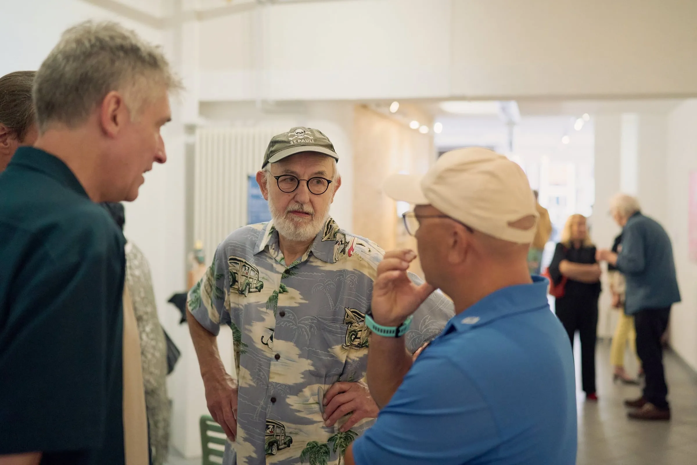 Drei Männer stehen im Gespräch im Ausstellungsraum, im Hintergrund weitere Gäste an hellen Wänden