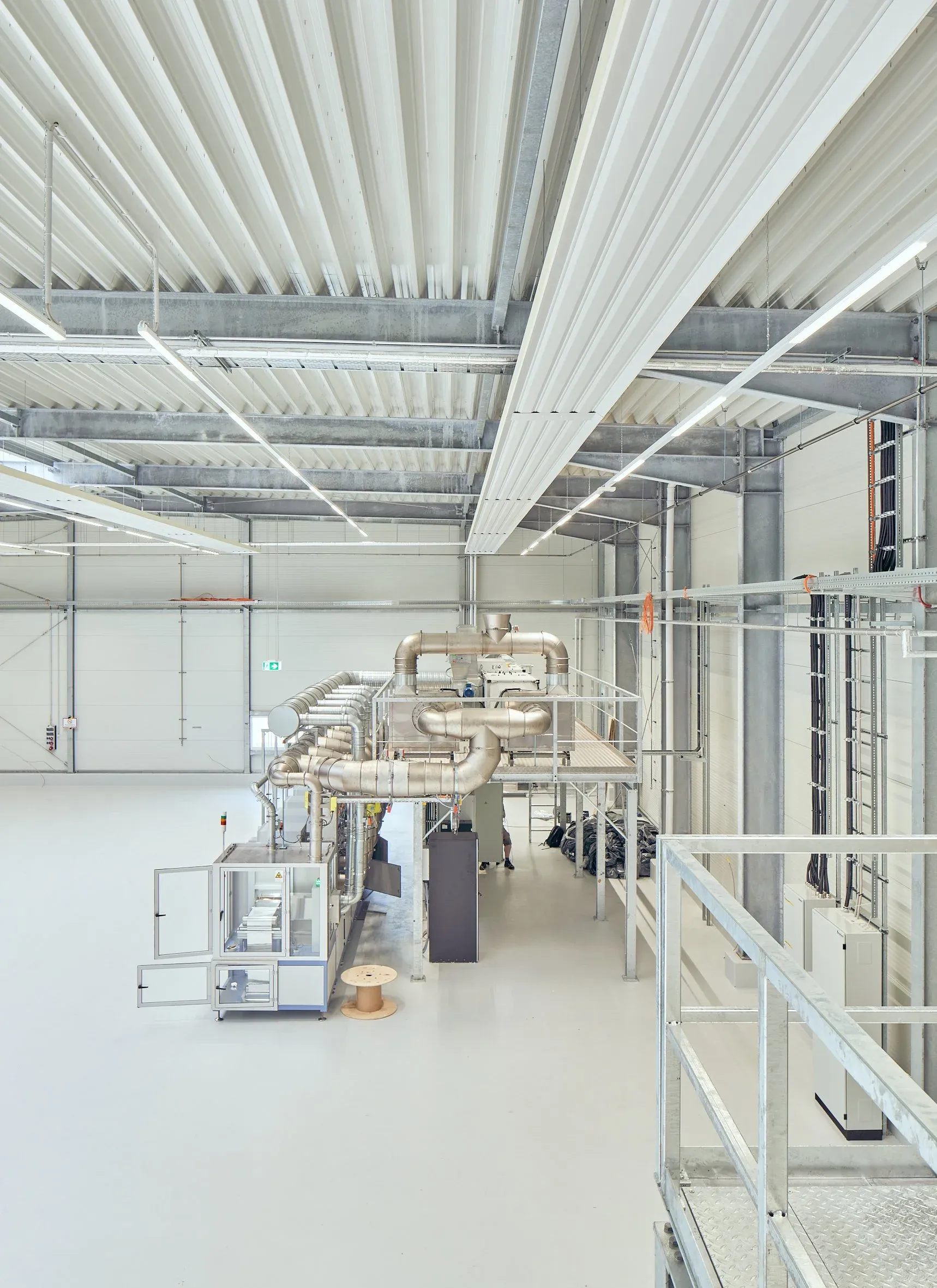 Blick von der Empore in die Halle mit Lüftungsrohren, Maschinen und sichtbarer Trapezblechdecke, helles Tageslicht