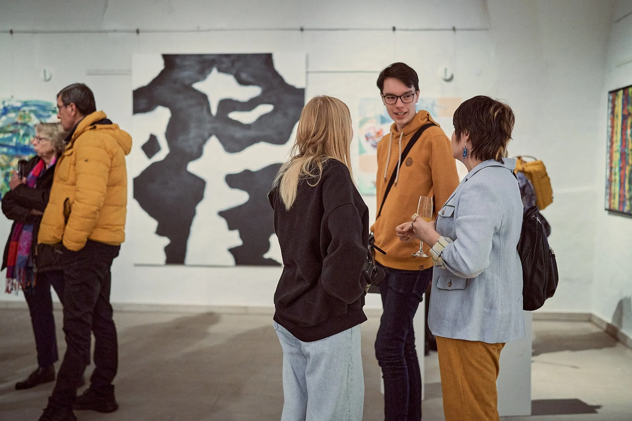 Vier Gäste stehen im Gespräch in der hellen Halle, an den Wänden ein großes schwarzweißes Werk und farbige Bilder
