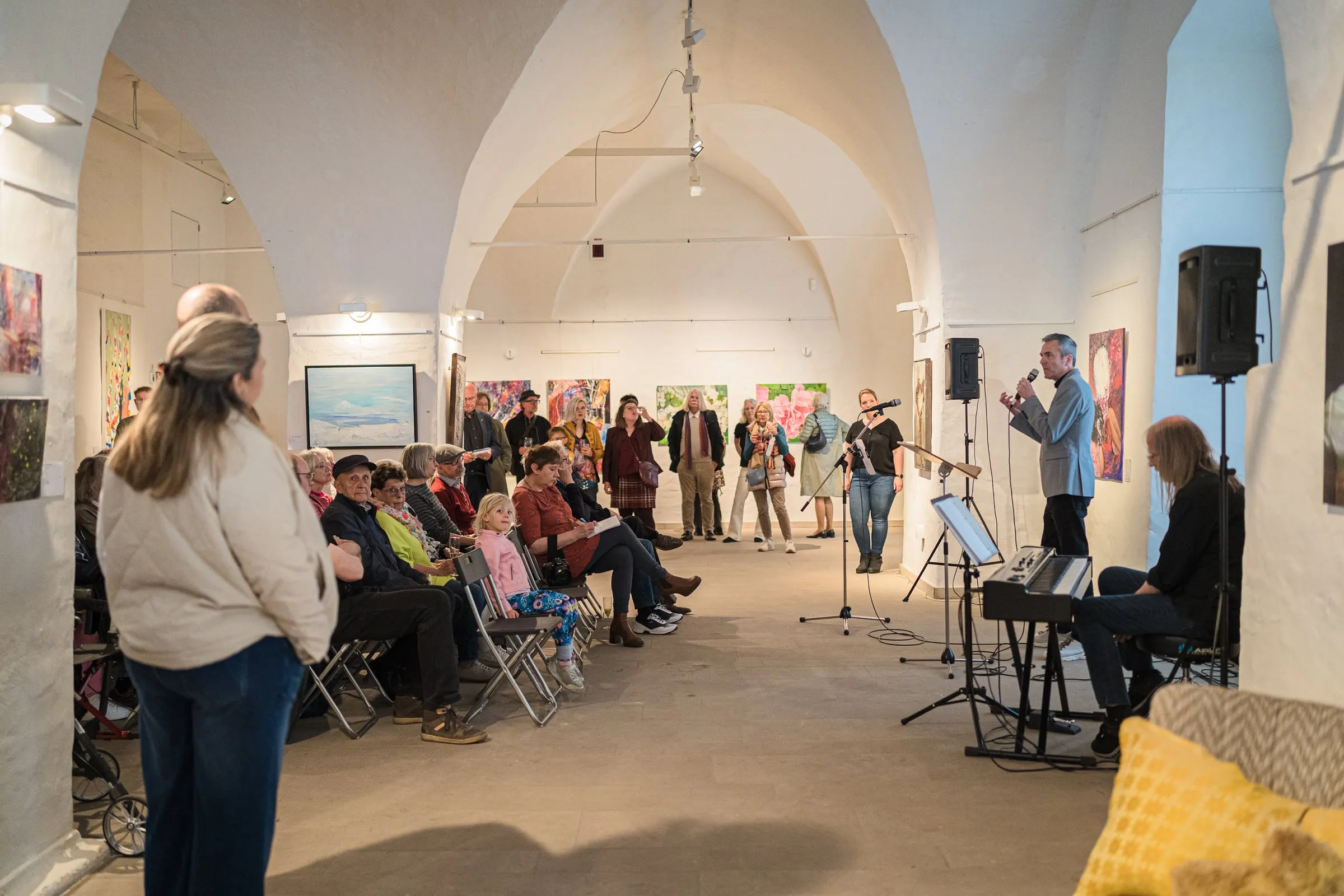 Eröffnung mit sitzendem Publikum, ein Redner am Mikrofon vor gehängten Werken im gewölbten Galerieraum