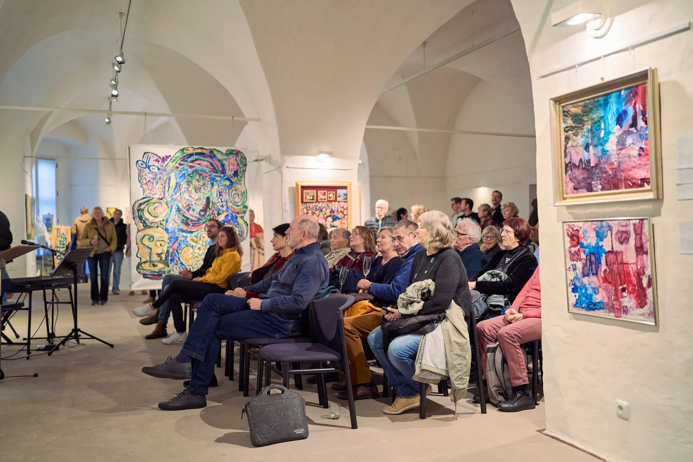 Sitzendes Publikum bei einer Eröffnung im gewölbten Saal, rechts gerahmte abstrakte Werke