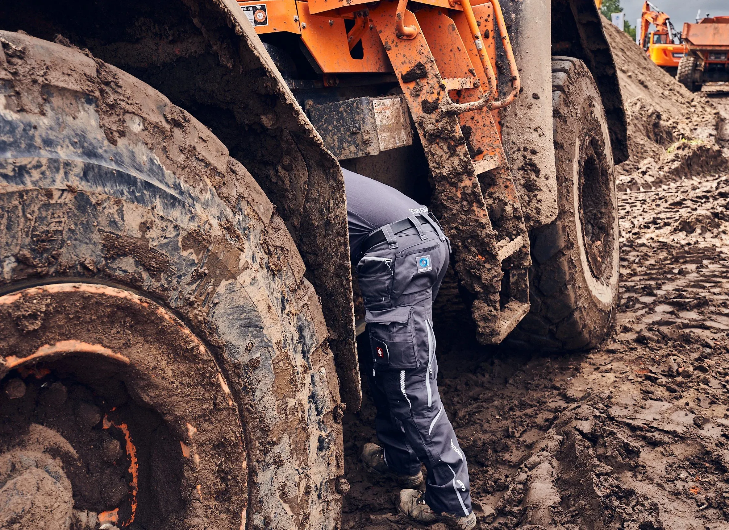 Person in Arbeitskleidung beugt sich zwischen die schlammigen Räder einer Baumaschine, Detail
