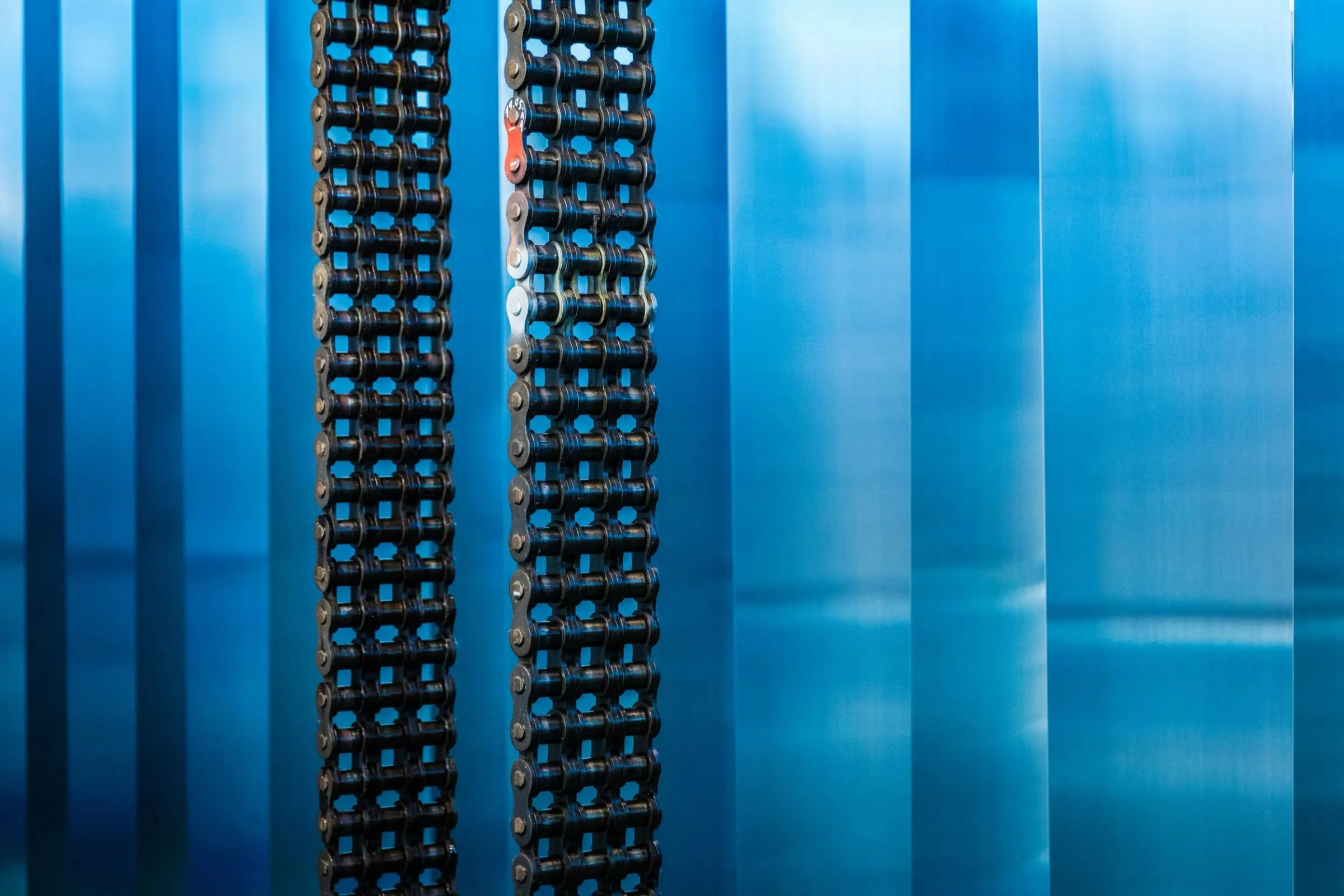 Detail einer Maschine bei Rheinzink, zwei dunkle Antriebsketten vor blau schimmernden, glänzenden Zinkbahnen