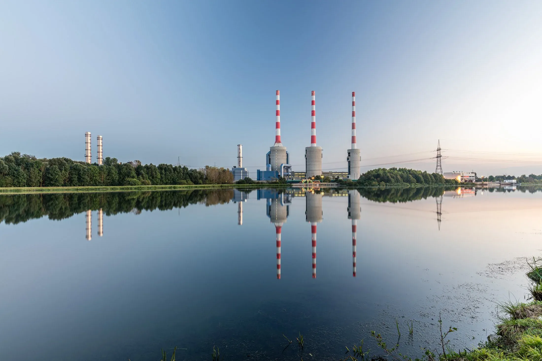 Industrieanlage am See, drei rotweiße Schornsteine und Hallen spiegeln sich im ruhigen Wasser, Außenaufnahme