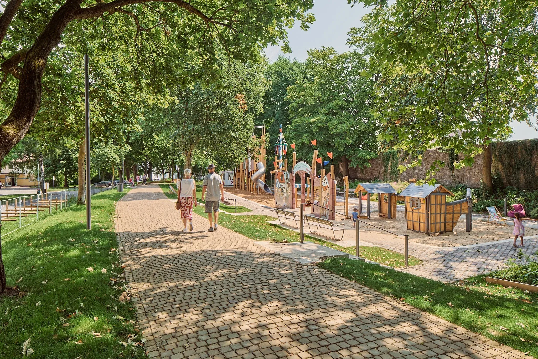 Gepflasterter Weg im Park führt zu einem Spielplatz aus Holz, ein Paar geht im Schatten der Bäume