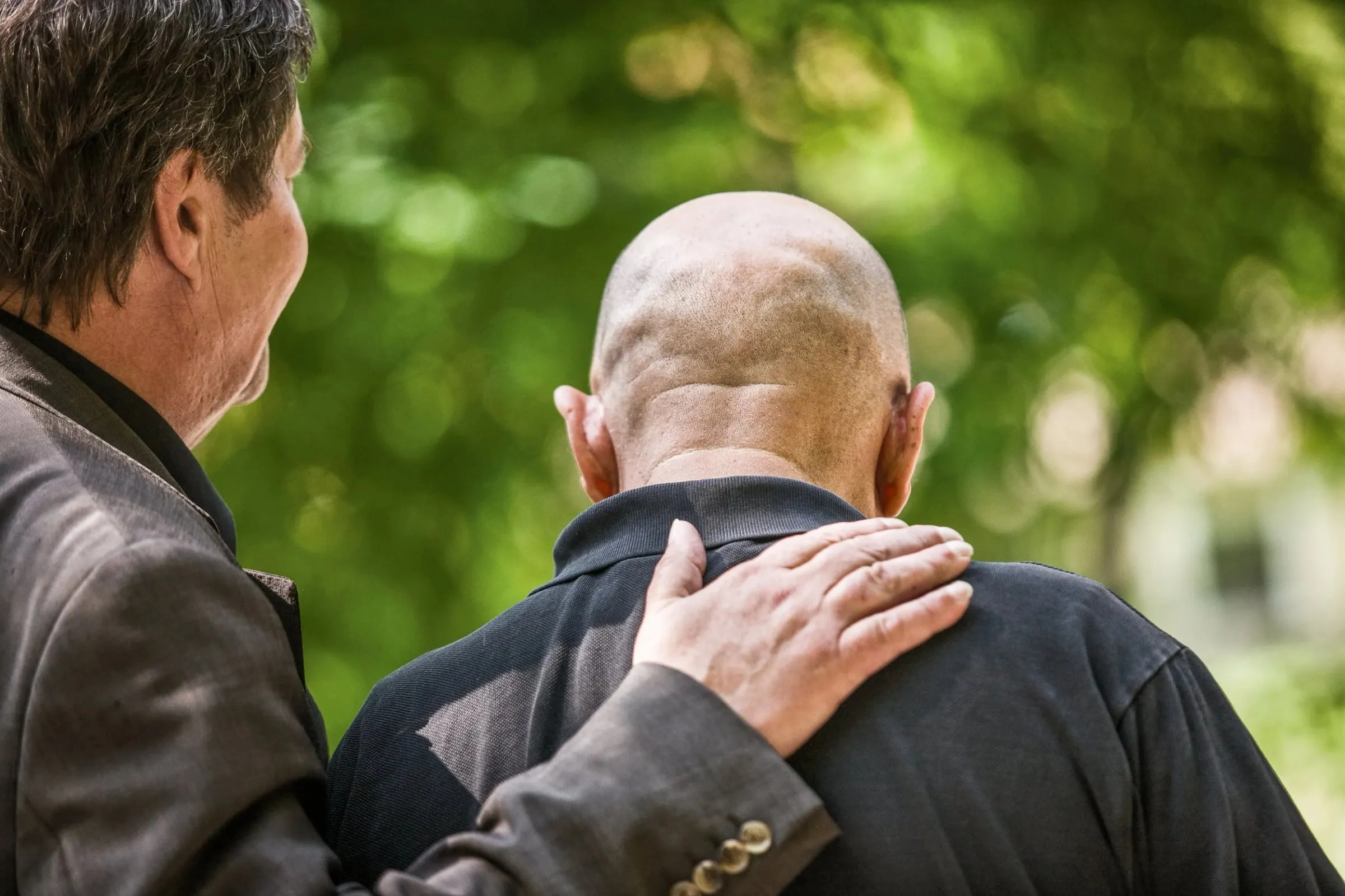 Zwei Männer von hinten, einer legt dem anderen die Hand auf die Schulter, grüner unscharfer Hintergrund