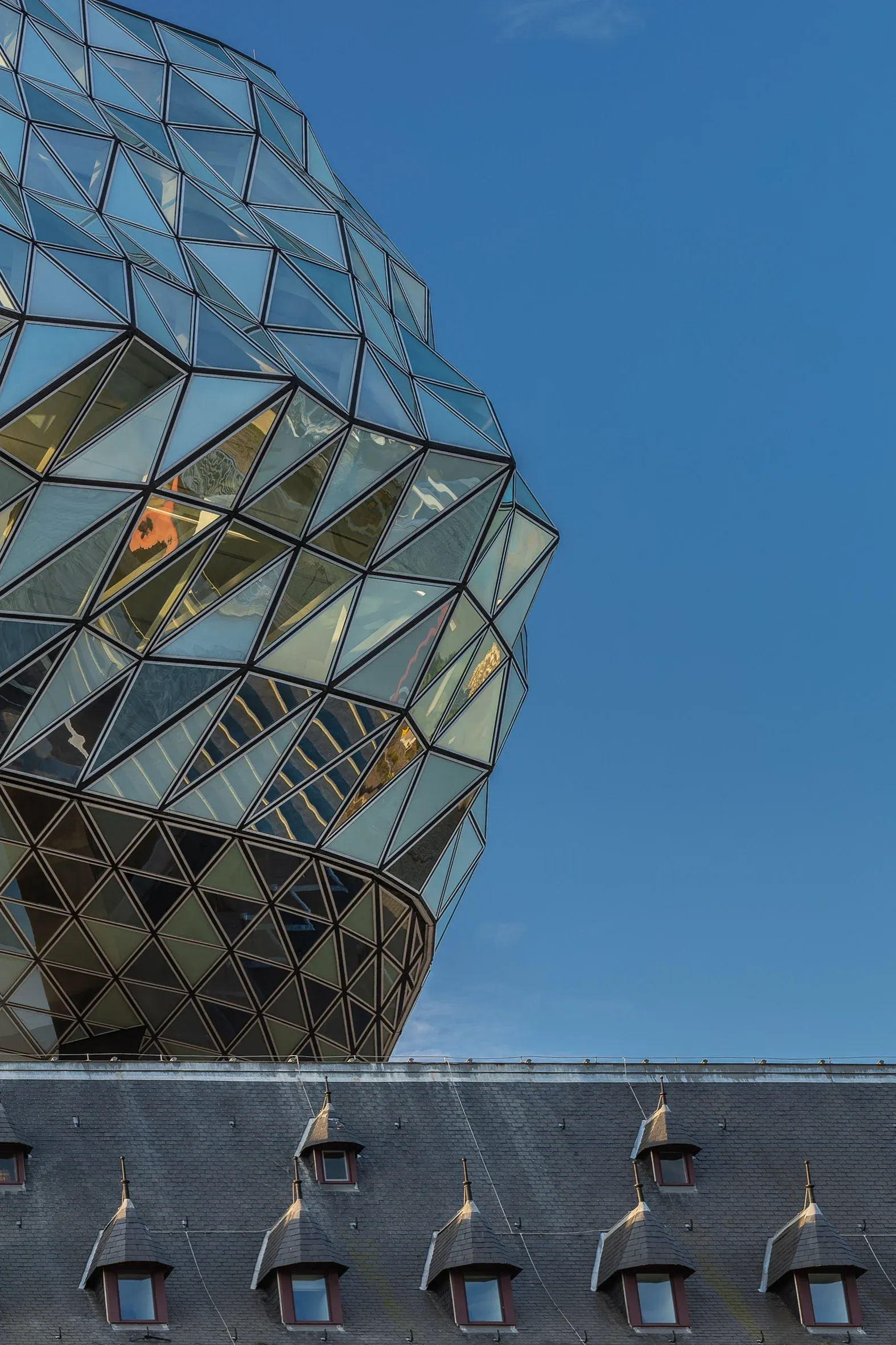 Detail von unten, die diagonal gerasterte Glasfassade des Aufbaus wölbt sich über dem Schieferdach mit Gauben des Altbaus