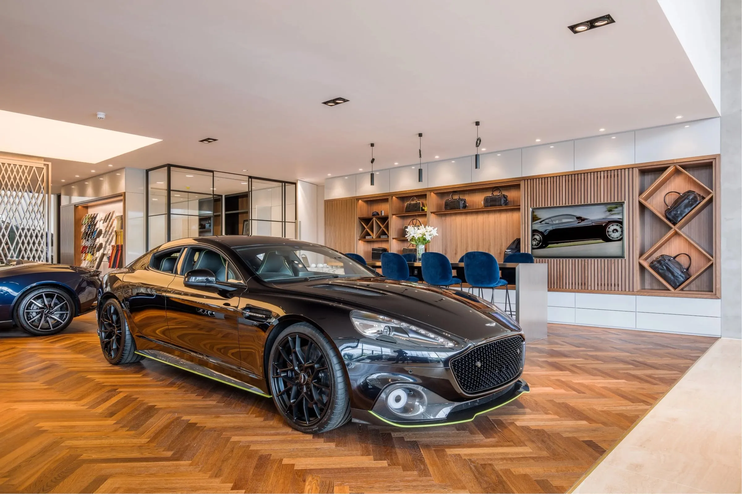 Innenraum mit schwarzem Aston Martin Sportwagen auf Parkett, im Hintergrund Lounge mit Vitrinen und blauen Sitzen