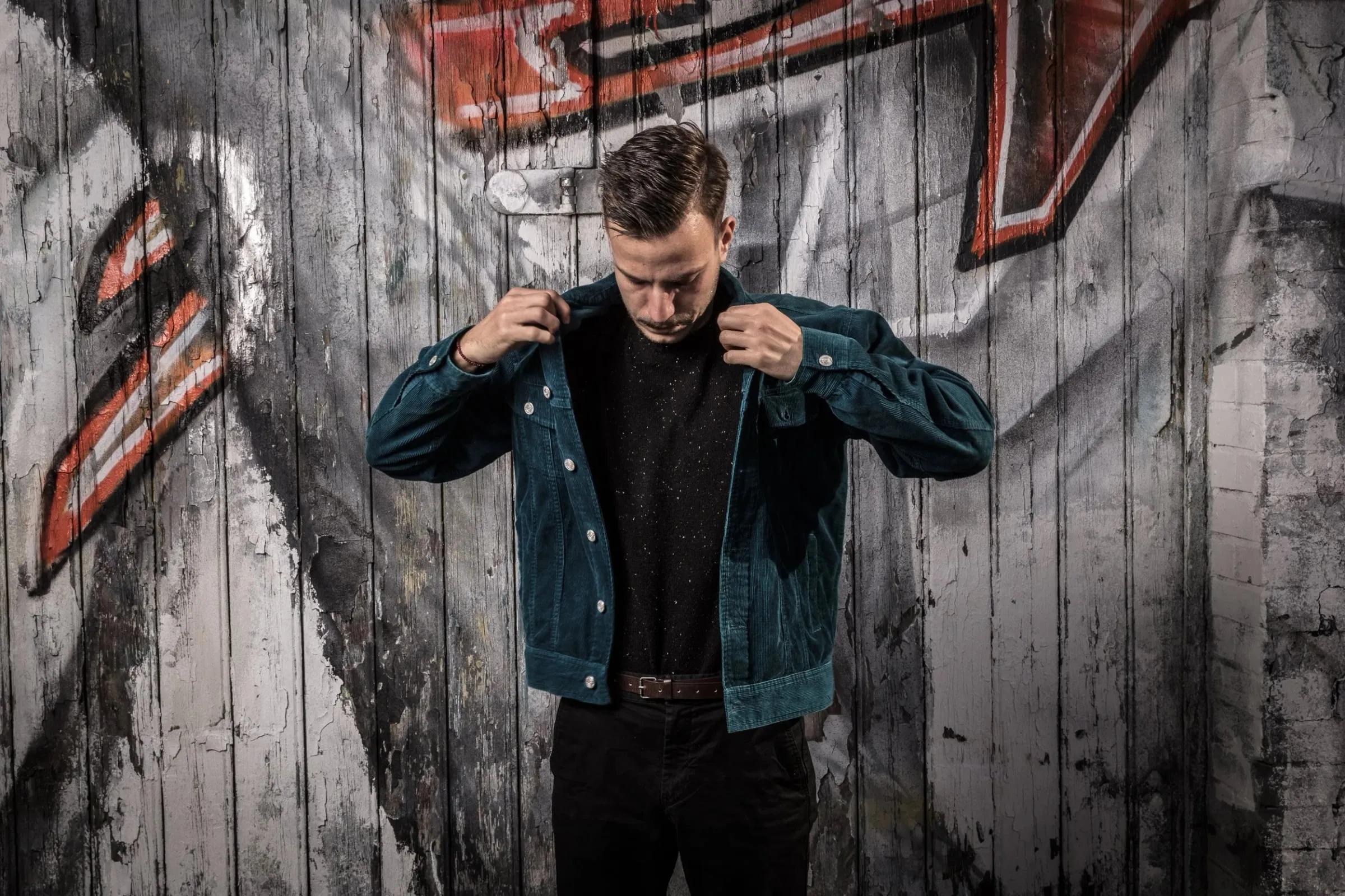 Junger Mann in dunkler Jeansjacke vor einer Holzwand mit Graffiti, Blick nach unten, greift an seinen Kragen, halbe Figur