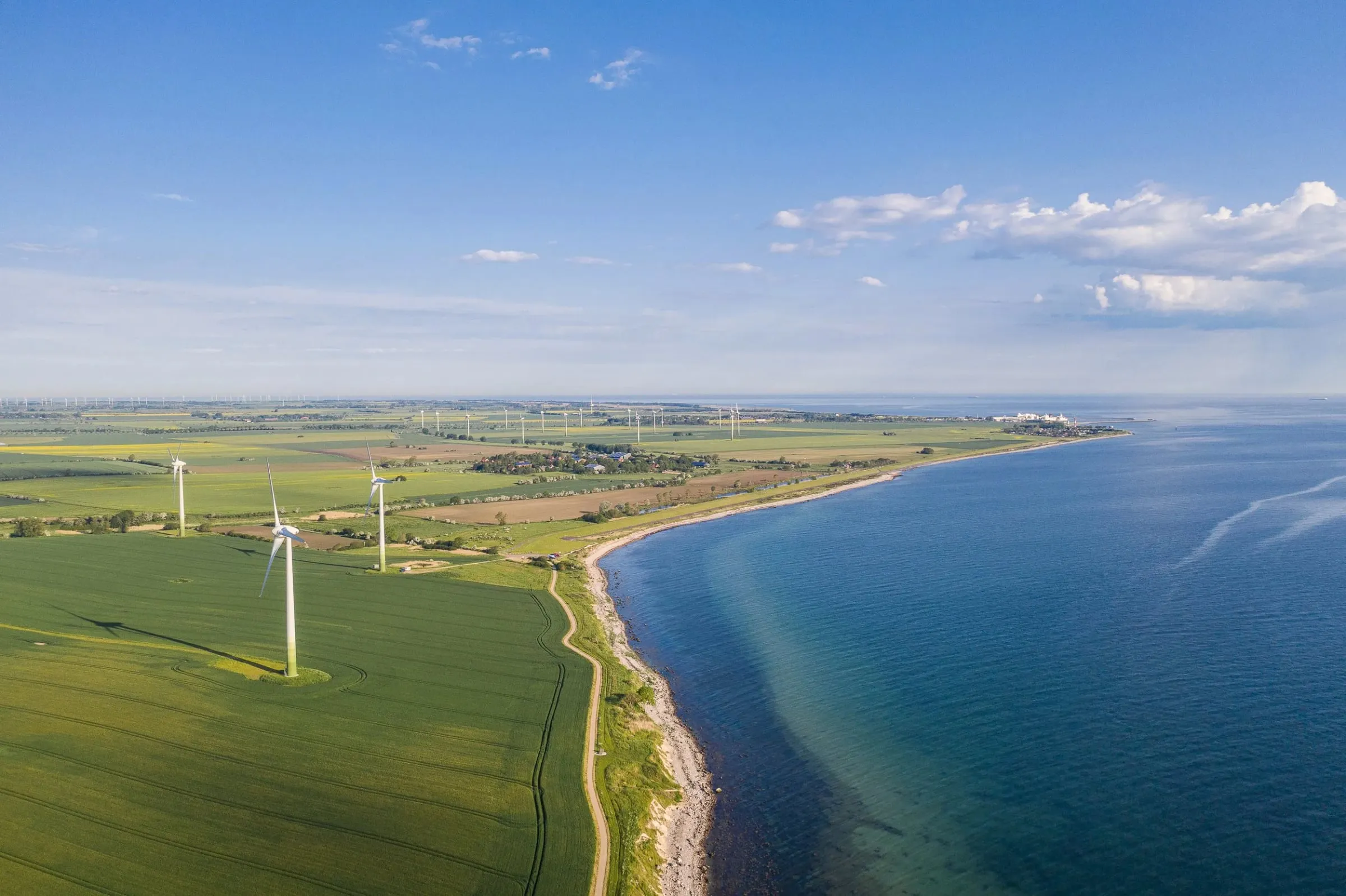 Luftaufnahme einer Küste mit Windrädern auf grünen Feldern entlang einer Kiesküste am Meer