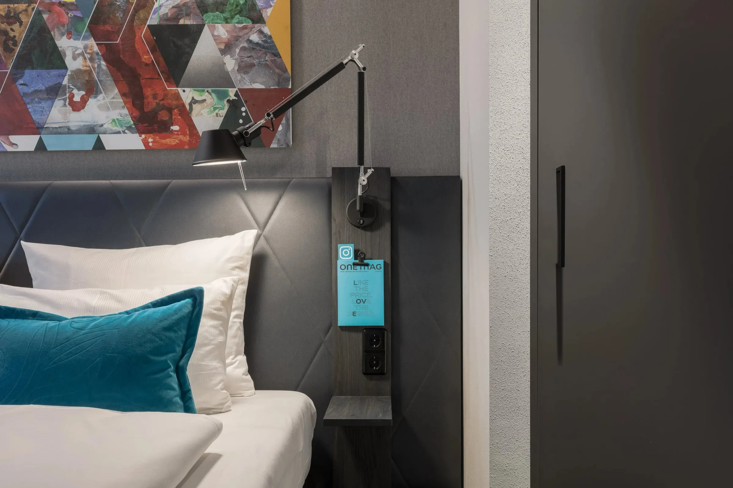 Detail eines Hotelzimmers, Bett mit weißer Wäsche und türkisem Kissen, schwarze Leselampe und abstraktes Wandbild über dem Kopfteil