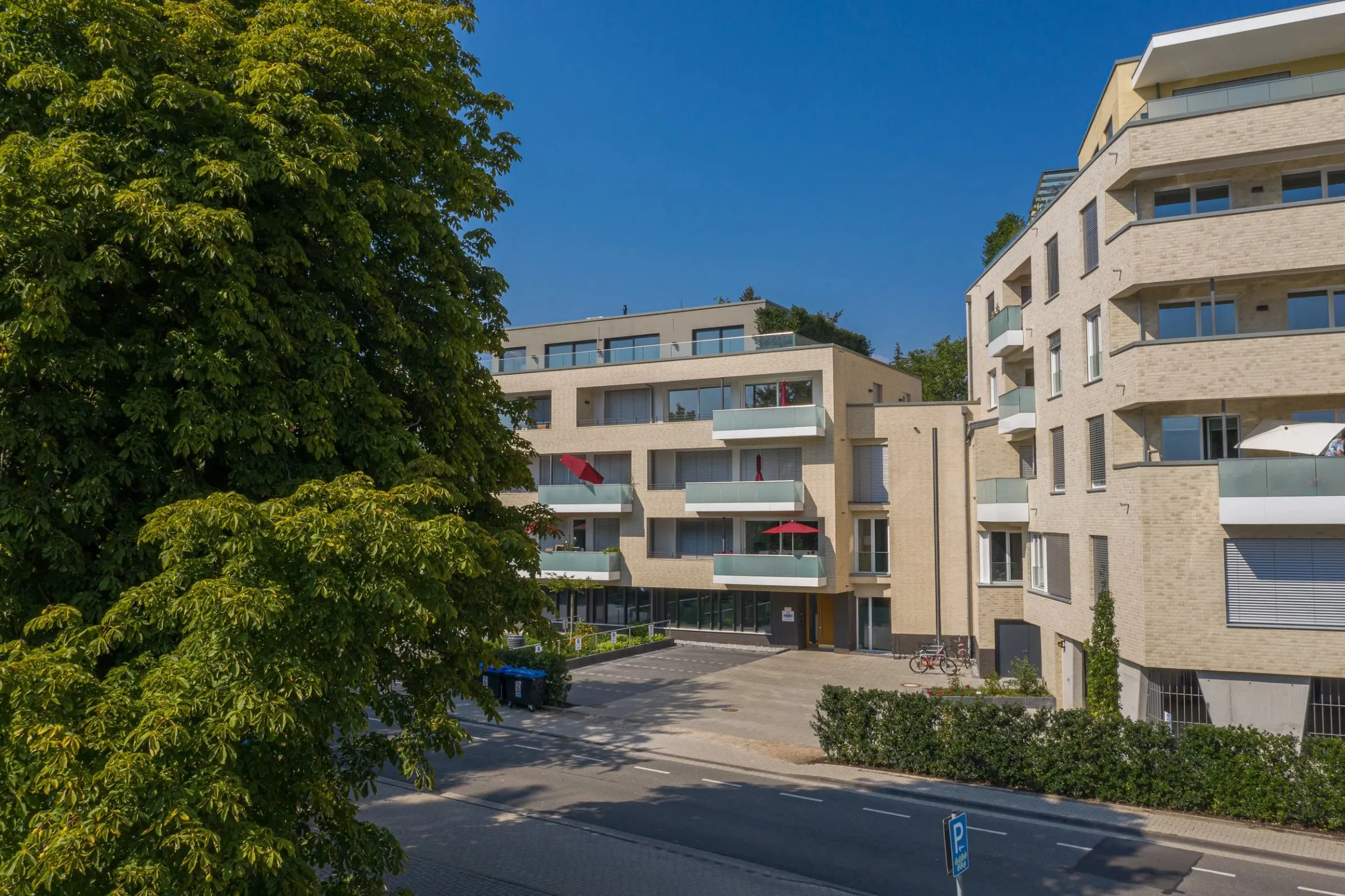 Luftaufnahme eines Wohnkomplexes mit heller Klinkerfassade und Balkonen, davor Straße und großer Baum bei Tageslicht