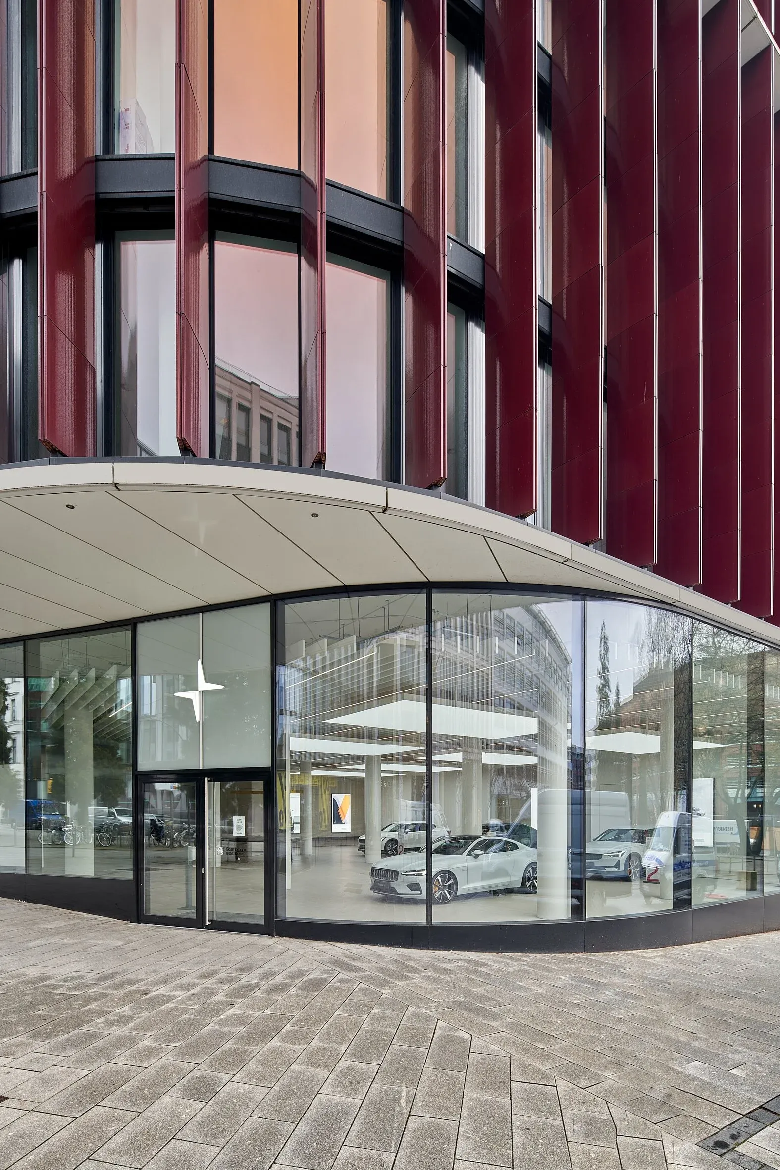 Außenansicht des Polestar Space, gerundete Glasecke unter roter Fassade, Fahrzeuge im Showroom sichtbar