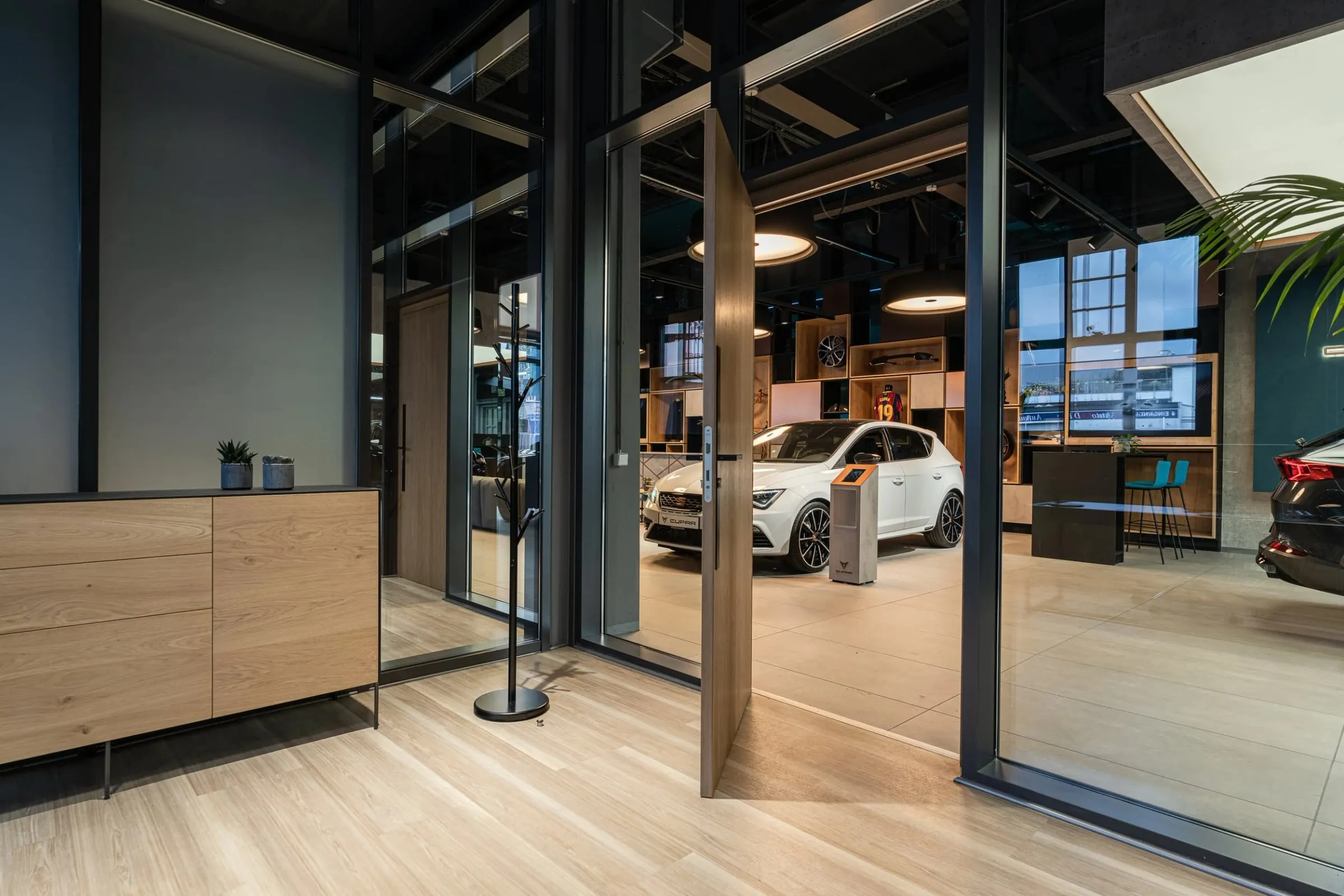 Blick durch offene Glastür auf weißen Cupra im Showroom, heller Holzboden und Sideboard im Vordergrund