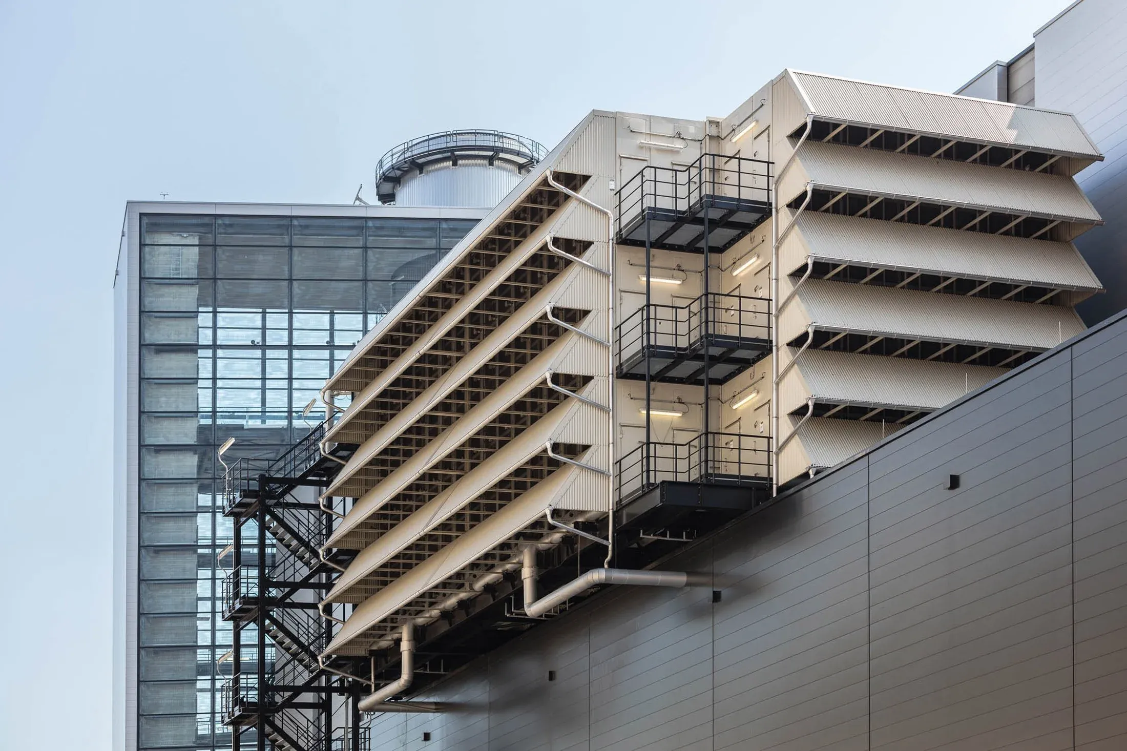 Außenfassade eines Industriegebäudes mit auskragenden Betonebenen, Stahltreppe und Glasfront vor hellem Himmel
