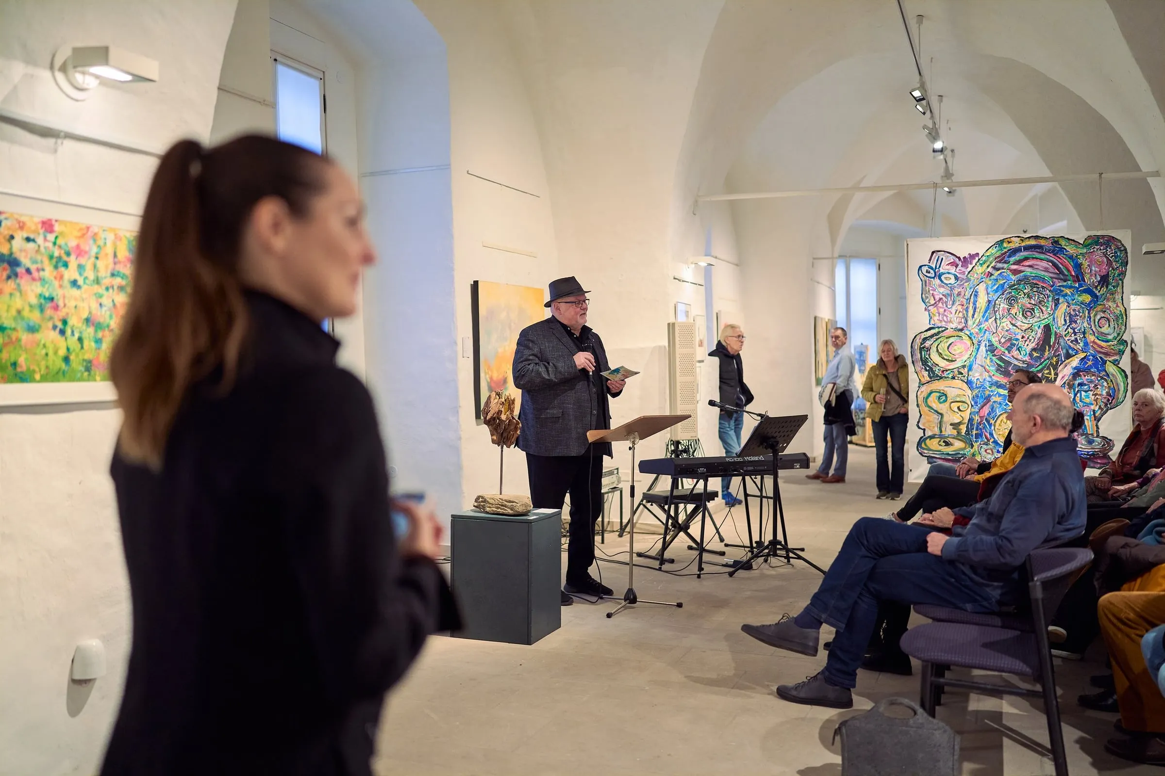 Mann hält eine Rede vor sitzendem Publikum im gewölbten Saal, ringsum farbige Werke