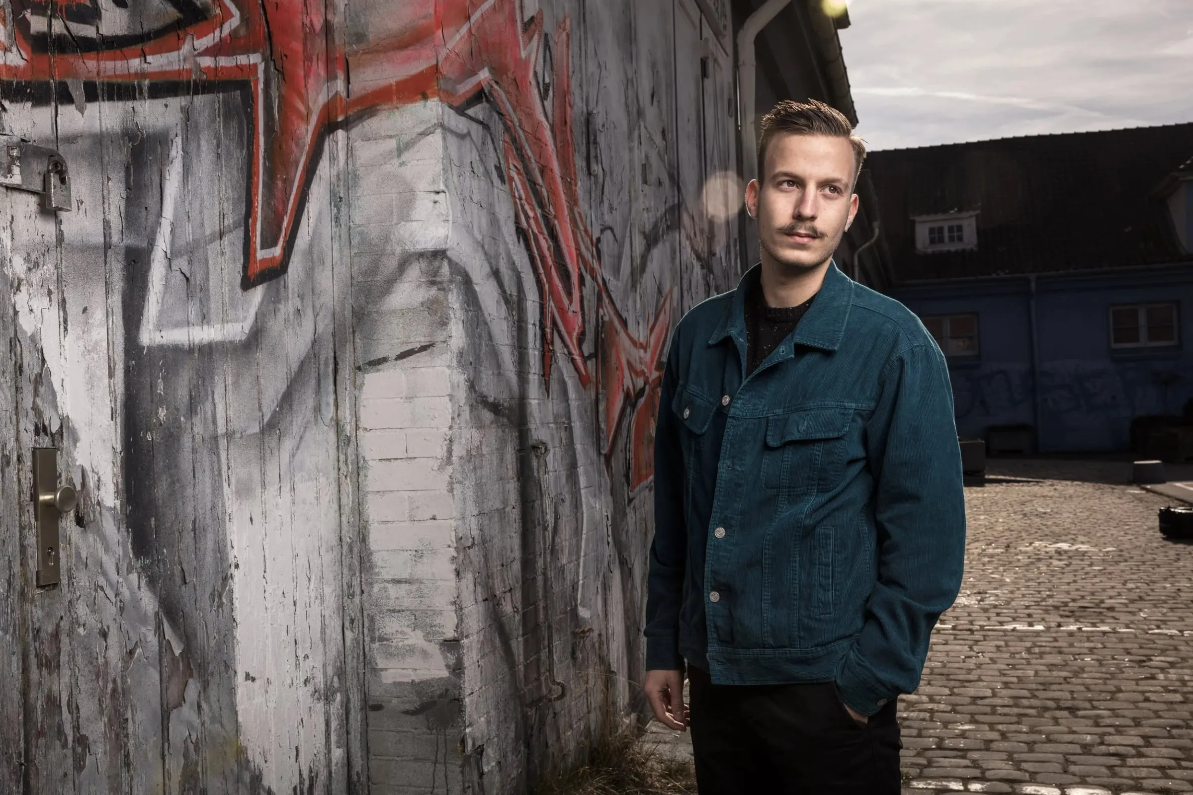 Junger Mann in dunkler Jeansjacke draußen neben einer Graffitiwand, Blick zur Seite, gepflasterter Hof im Hintergrund
