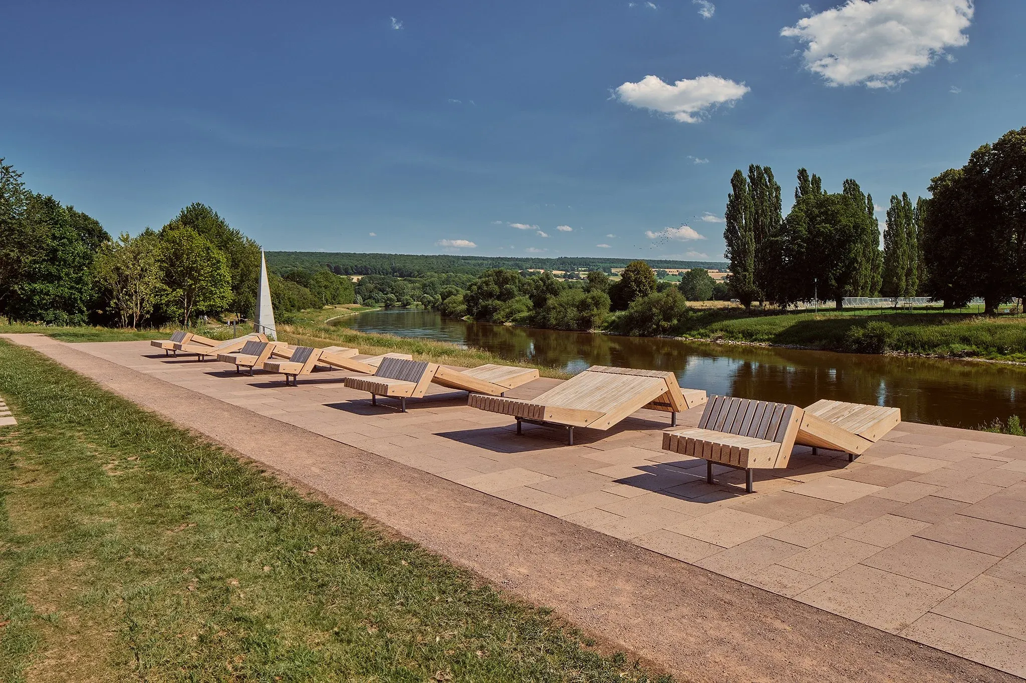 Reihe hölzerner Liegen auf einer gepflasterten Uferterrasse über der Weser, Landschaft und blauer Himmel