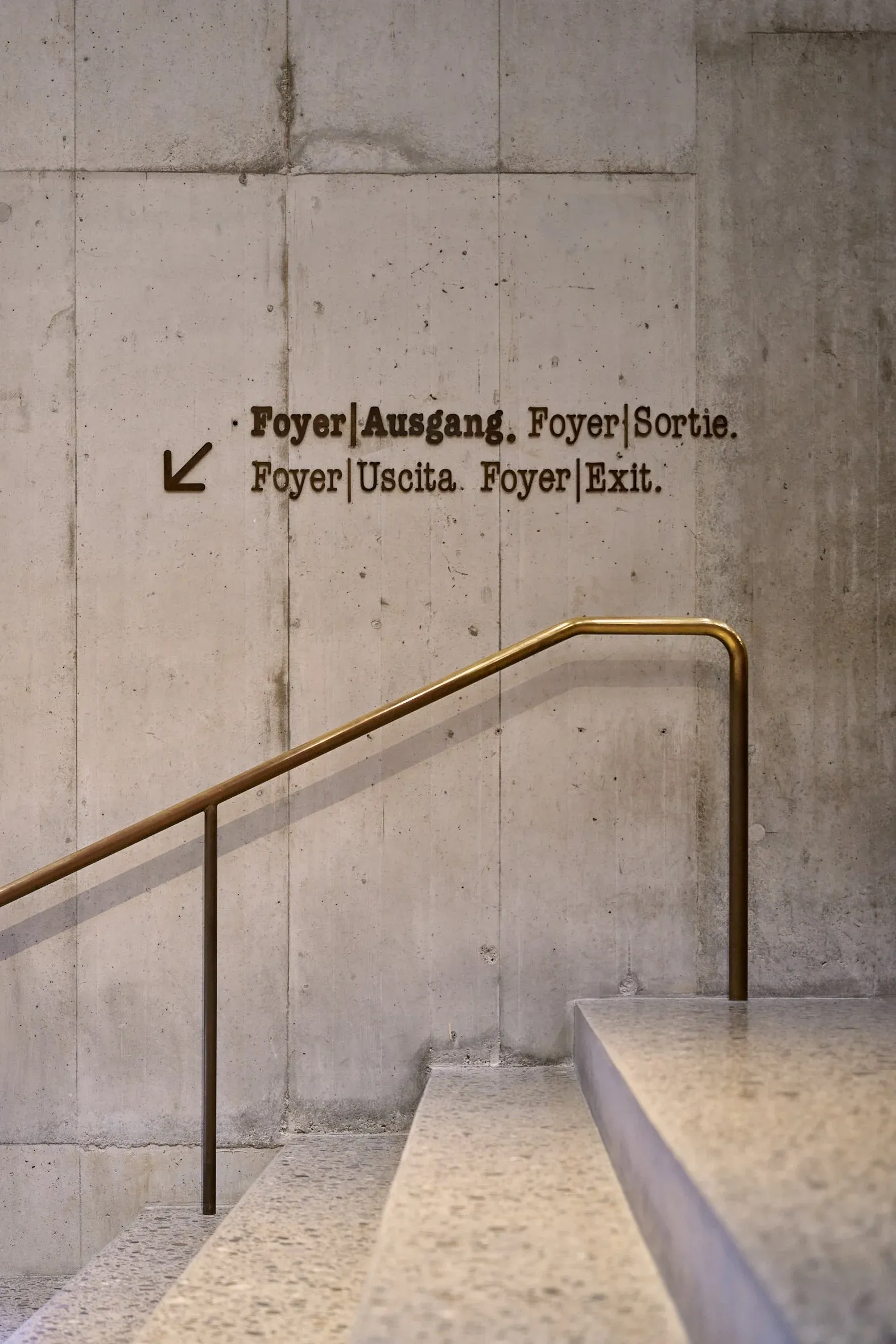 Sichtbetonwand mit mehrsprachiger Beschriftung Foyer und Ausgang und Messinggeländer über einer Treppe
