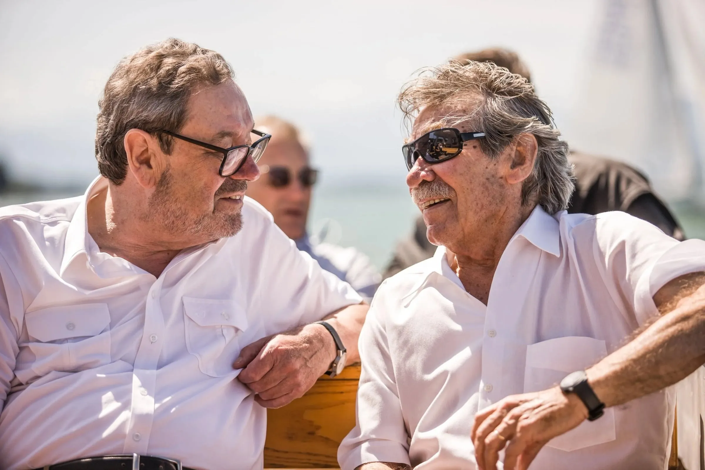 Zwei Männer im Gespräch im Sonnenlicht am Wasser, einer mit Brille zugewandt, im Hintergrund weitere Personen