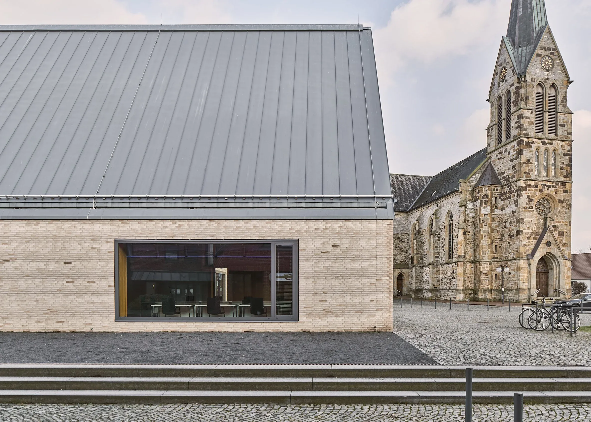 Außenaufnahme des Neubaus mit großem Panoramafenster neben der historischen Backsteinkirche von Bissendorf, gepflasterter Platz