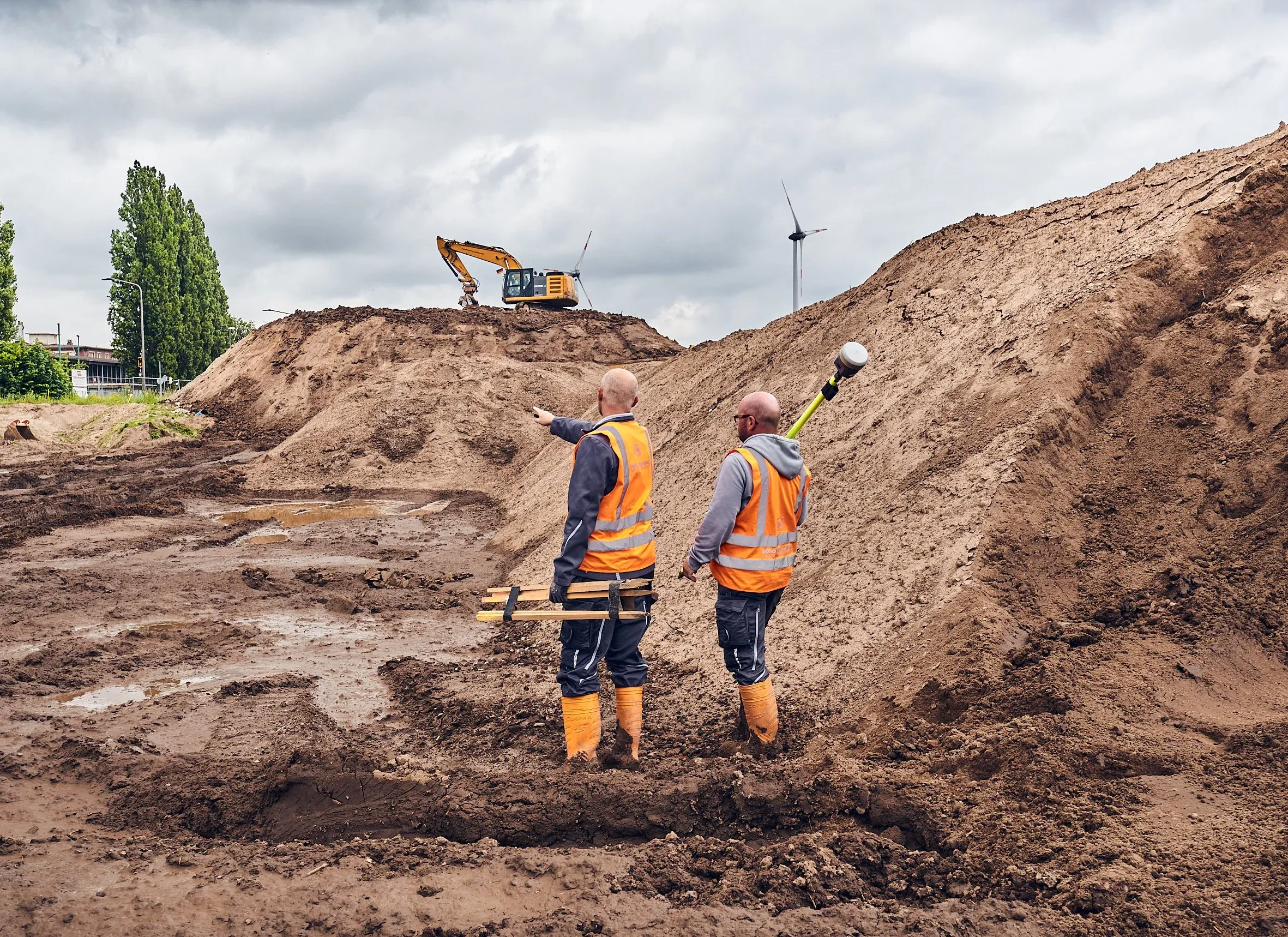 Zwei Arbeiter in Warnwesten vermessen am Sandhang, im Hintergrund ein Bagger und ein Windrad
