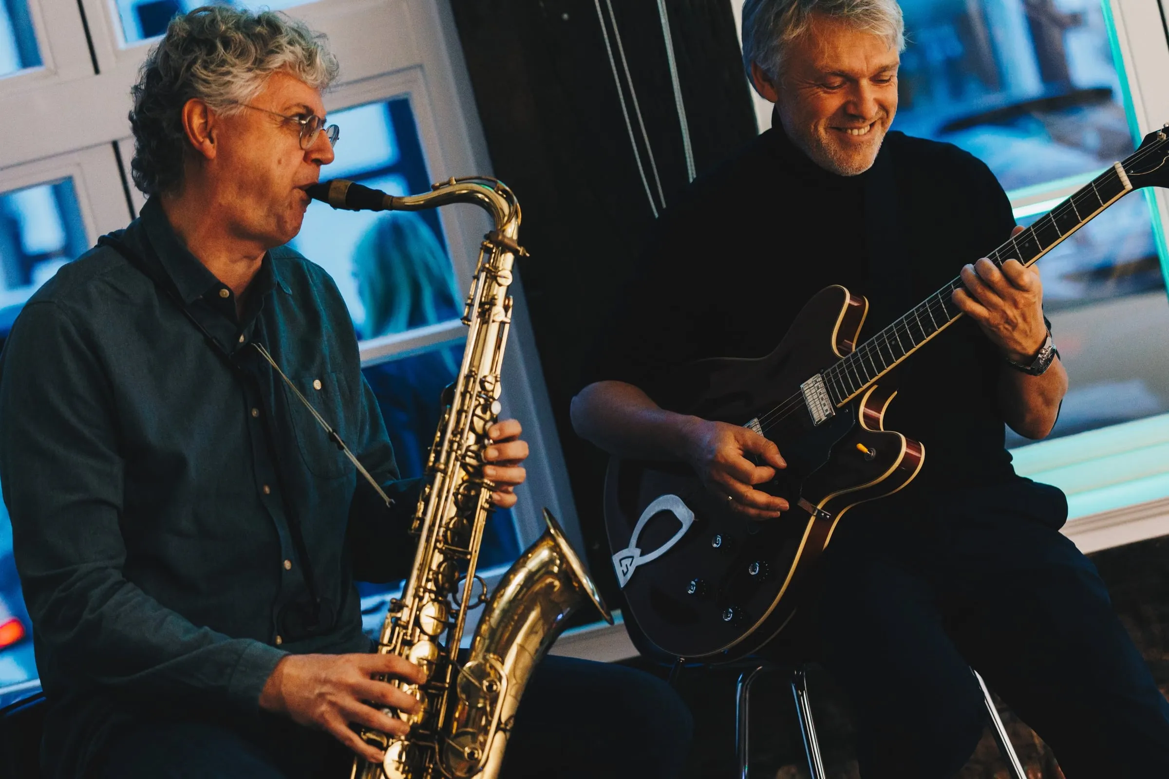 Zwei lächelnde Musiker mit Saxophon und Gitarre vor blau beleuchteten Fenstern