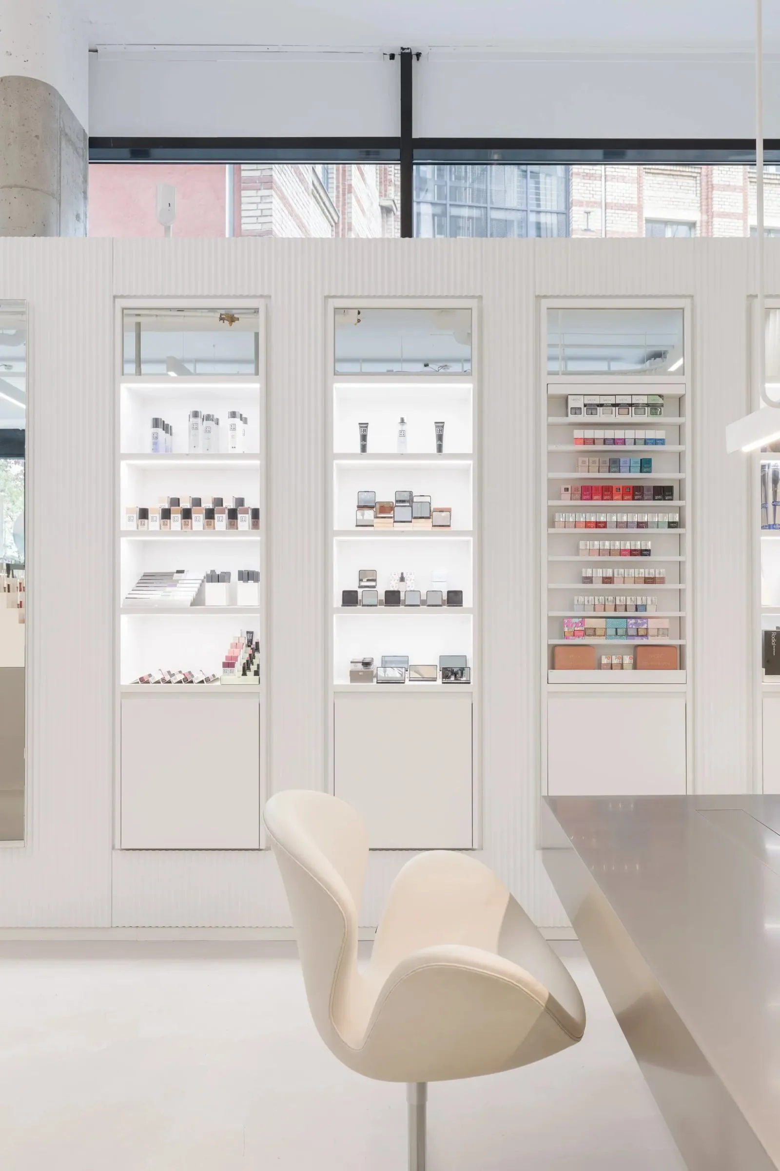 Beleuchtete weiße Vitrinen mit Kosmetik und Make-up, davor ein cremefarbener Designstuhl vor der Fensterfront