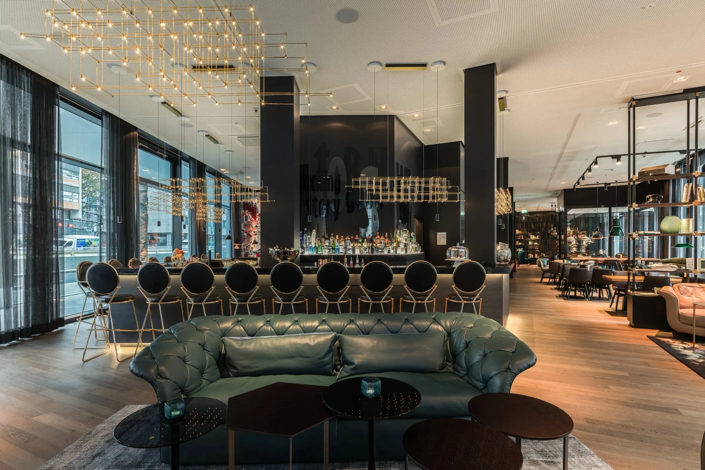 Bar und Lounge innen mit grünem Chesterfield-Sofa, Barhockern am Tresen und goldener Lichtinstallation an der Decke