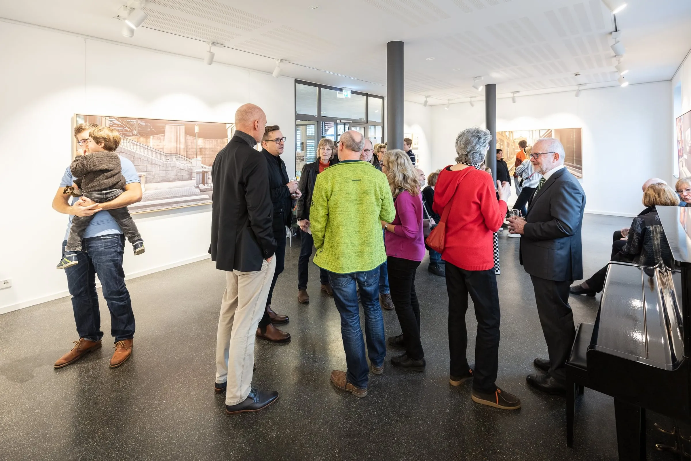 Besucher im Gespräch im Galerieraum, an den Wänden großformatige Fotografien, rechts ein Flügel