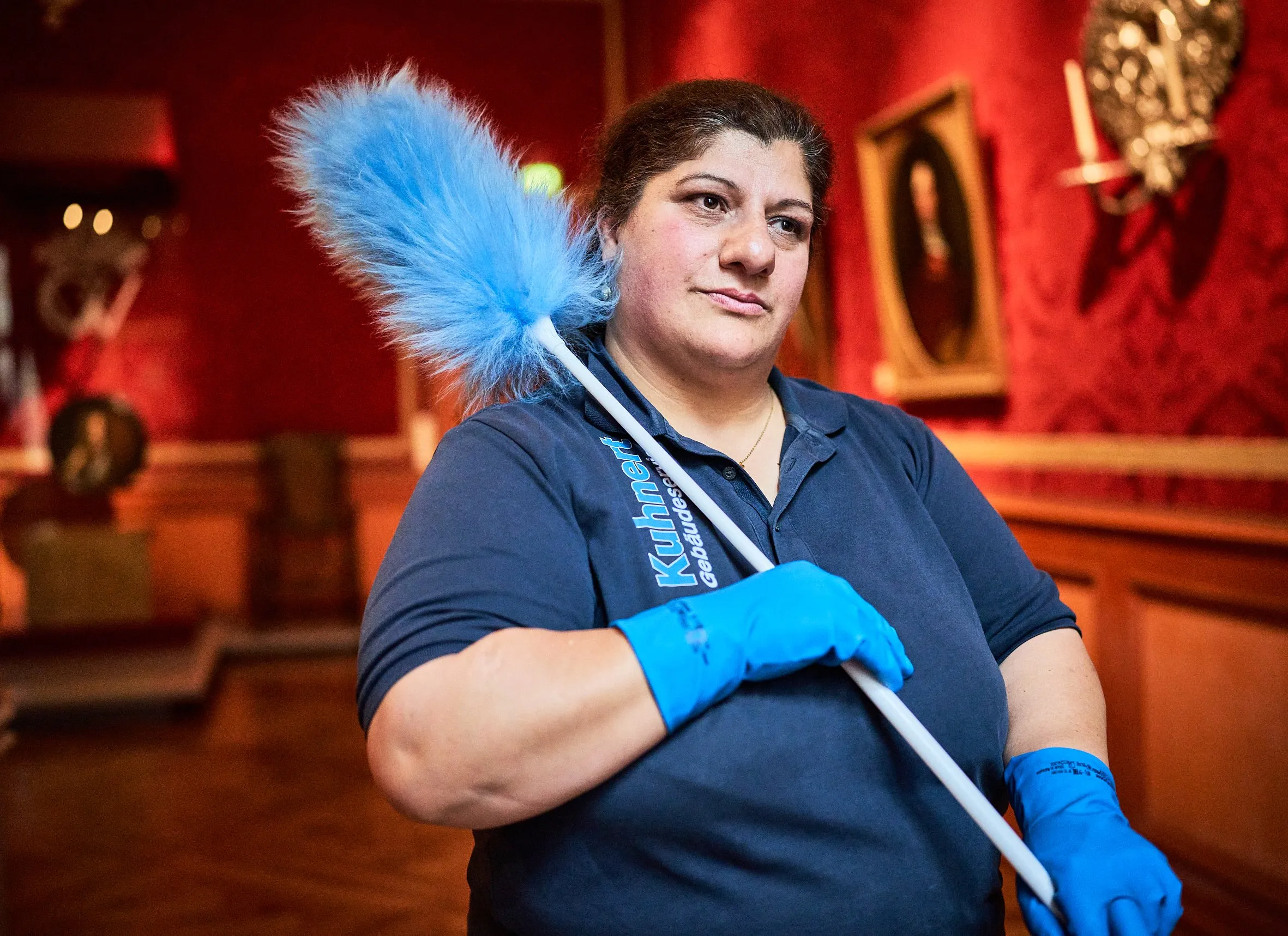 Porträt einer Mitarbeiterin im Kuhnert-Shirt mit blauem Staubwedel im roten Saal, blaue Handschuhe