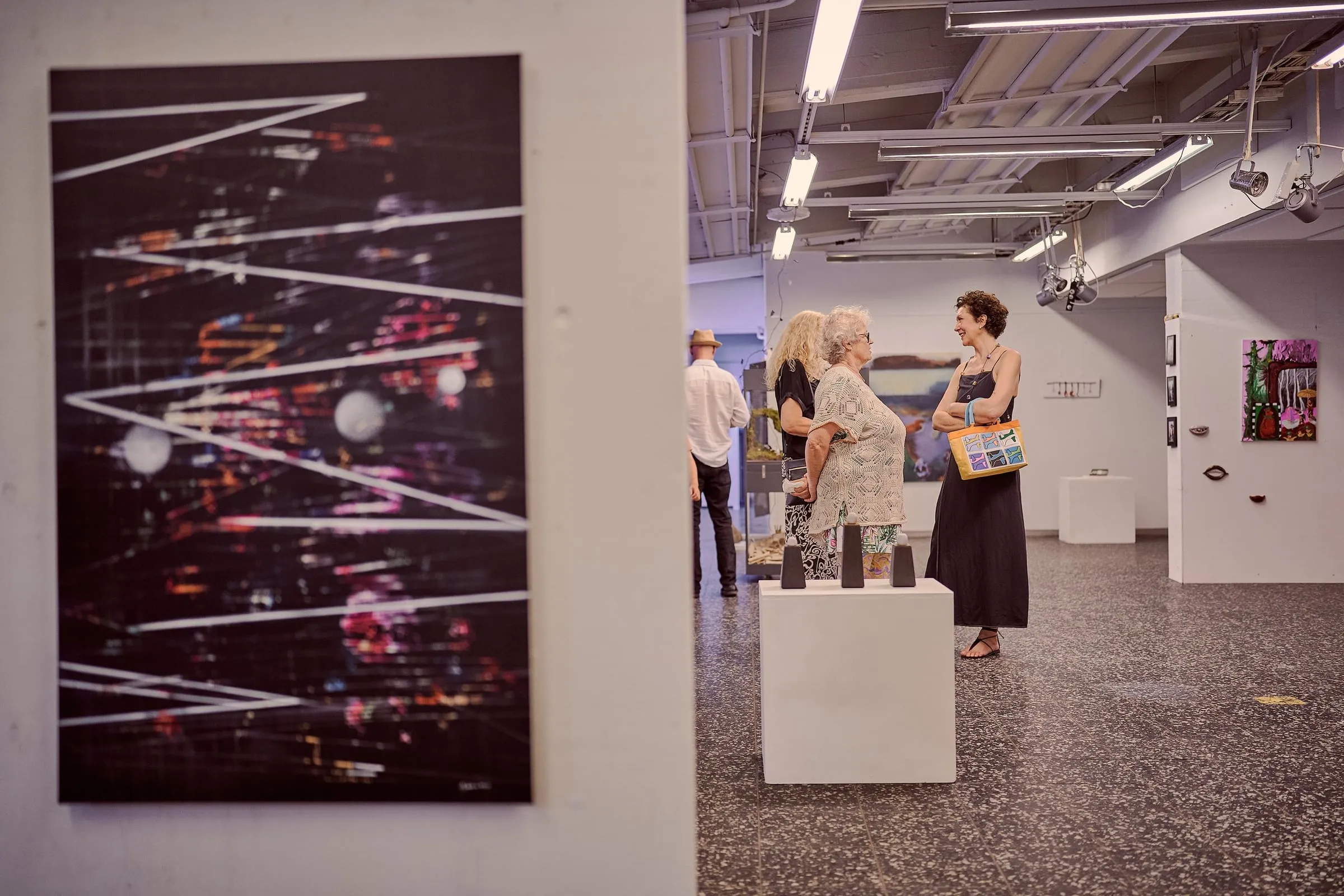 Links eine dunkle Fotoarbeit an der Wand, im Hintergrund Besucherinnen neben einem Sockel im Raum