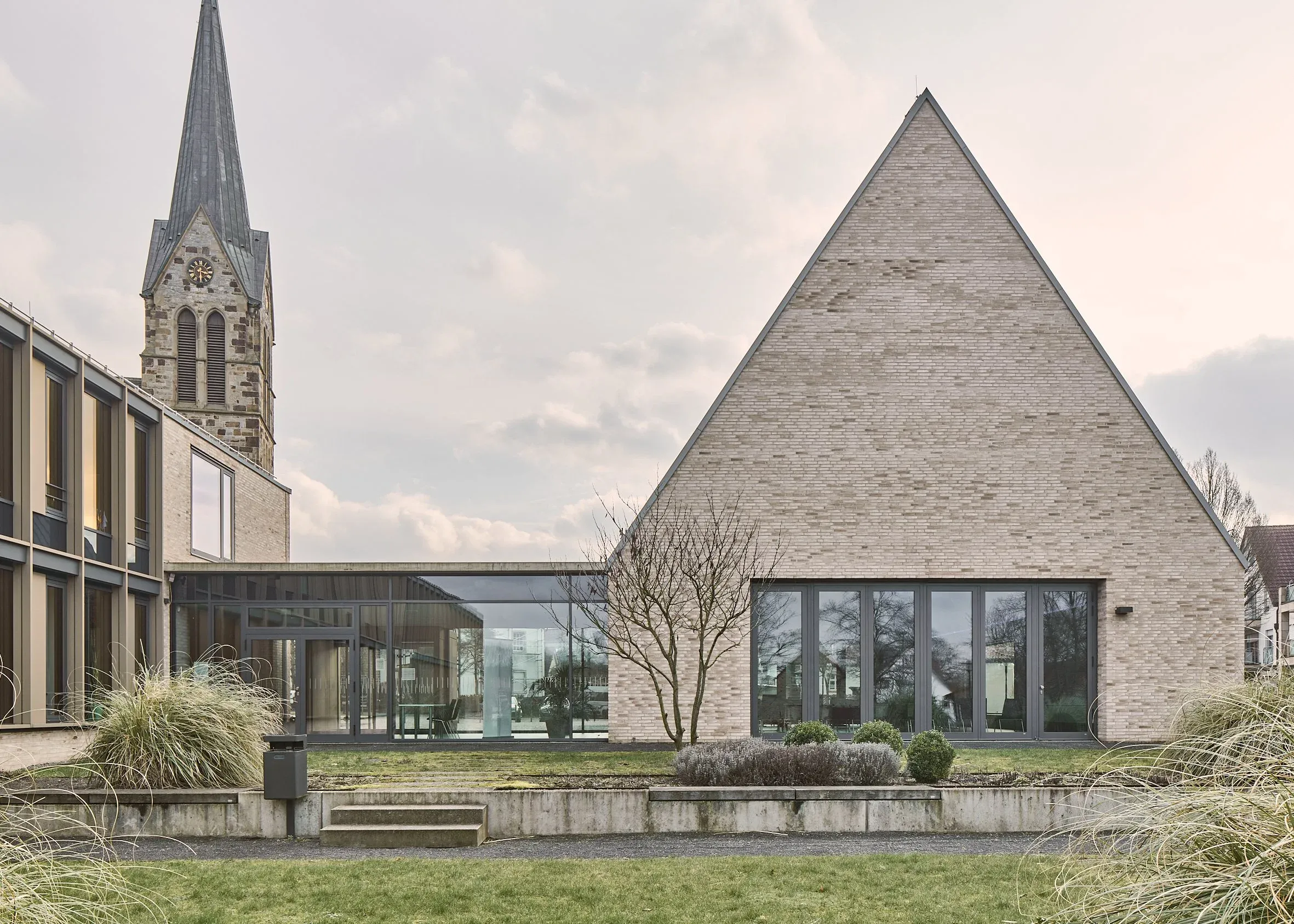 Außenansicht mit spitzem Klinkergiebel, verglastem Verbindungsbau und Kirchturm im Hintergrund, davor Rasen und kahler Baum