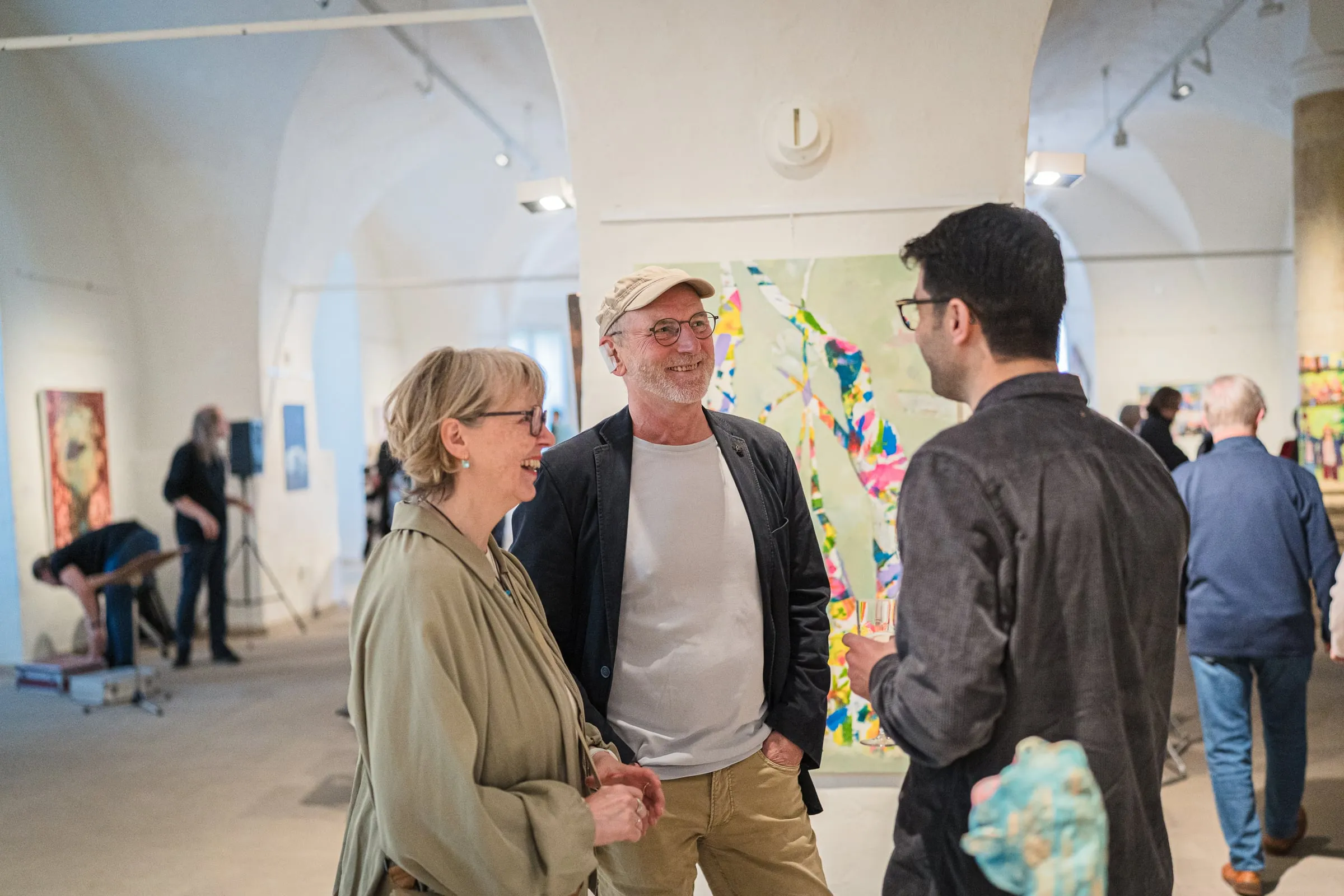 Drei Besucher im Gespräch im gewölbten Galerieraum, an den Wänden gehängte Werke, weitere Gäste im Hintergrund