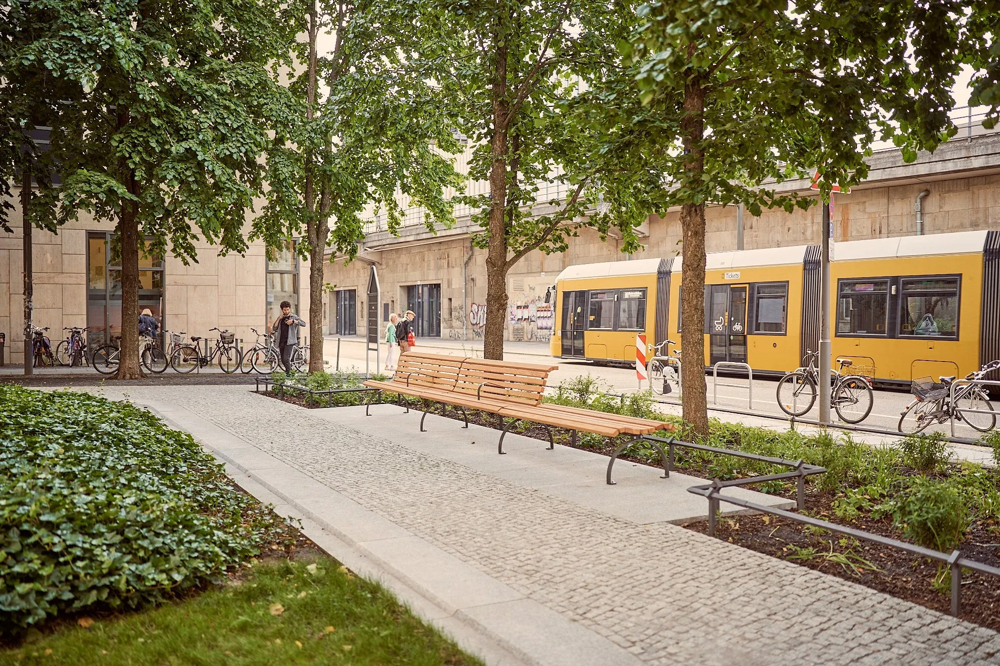 Lange Holzbank und gepflasterter Weg am Platz, im Hintergrund gelbe Straßenbahn und Fahrradständer