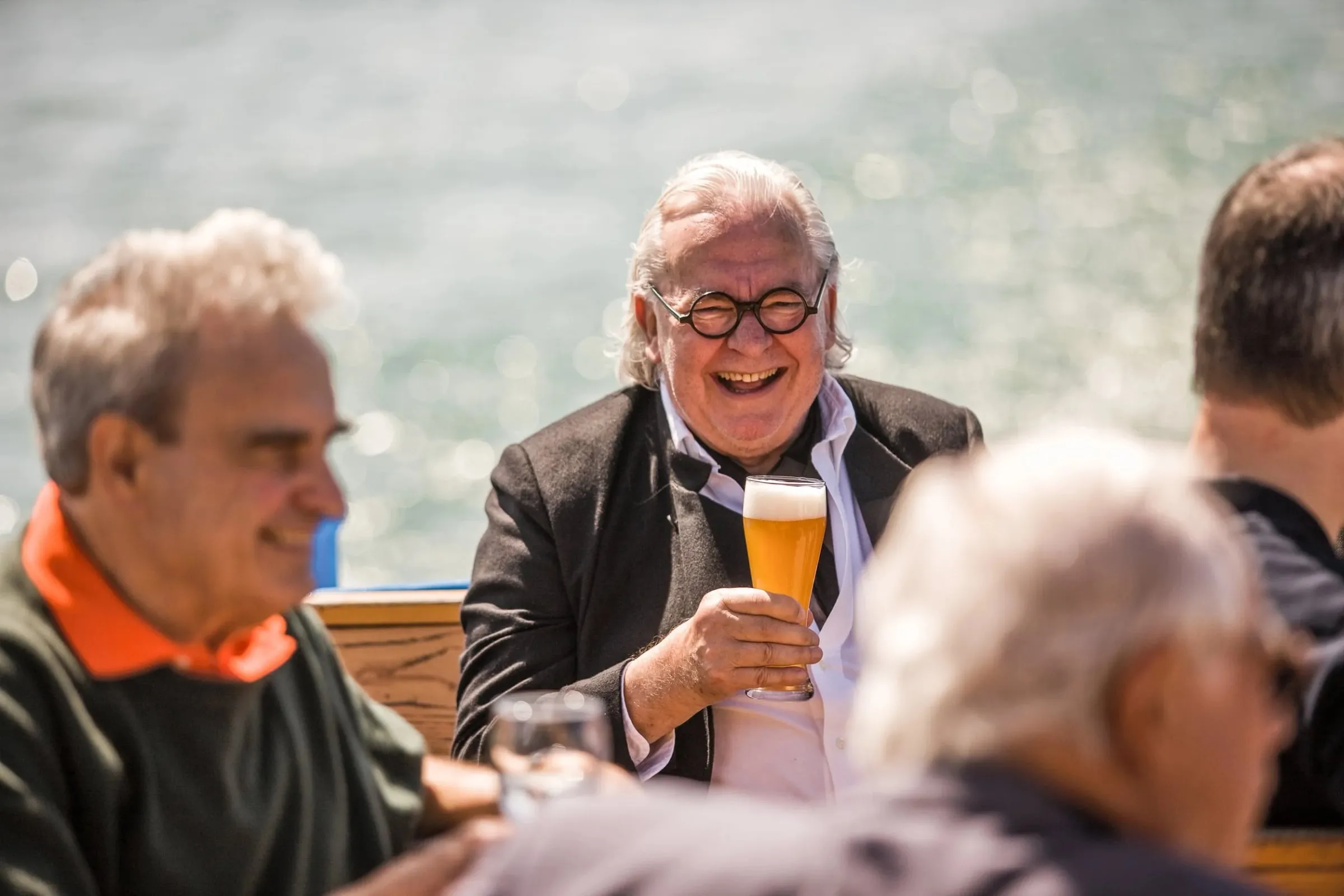 Mann mit Brille lacht und hält ein Glas, an einem Tisch im Freien am Wasser, weitere Personen ringsum