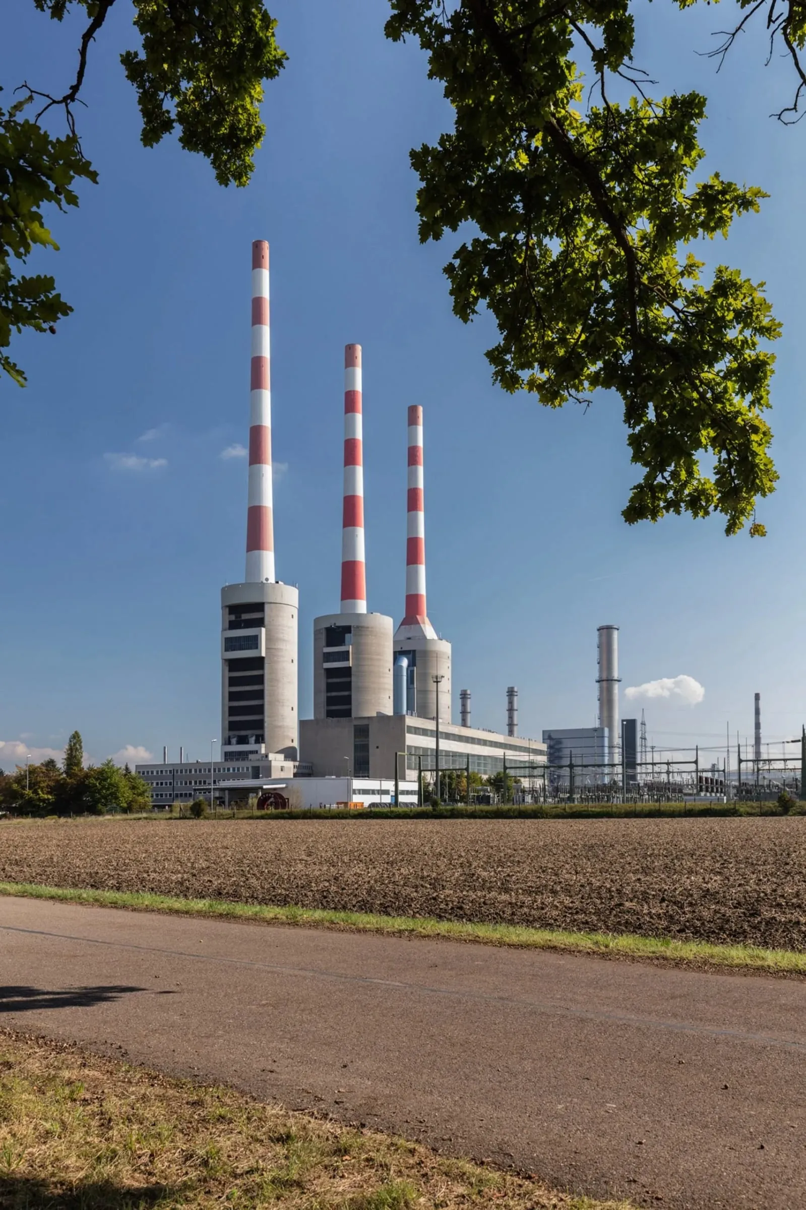 Industriewerk mit drei hohen rotweißen Schornsteinen hinter einem Acker, gerahmt von Laub, Tageslicht