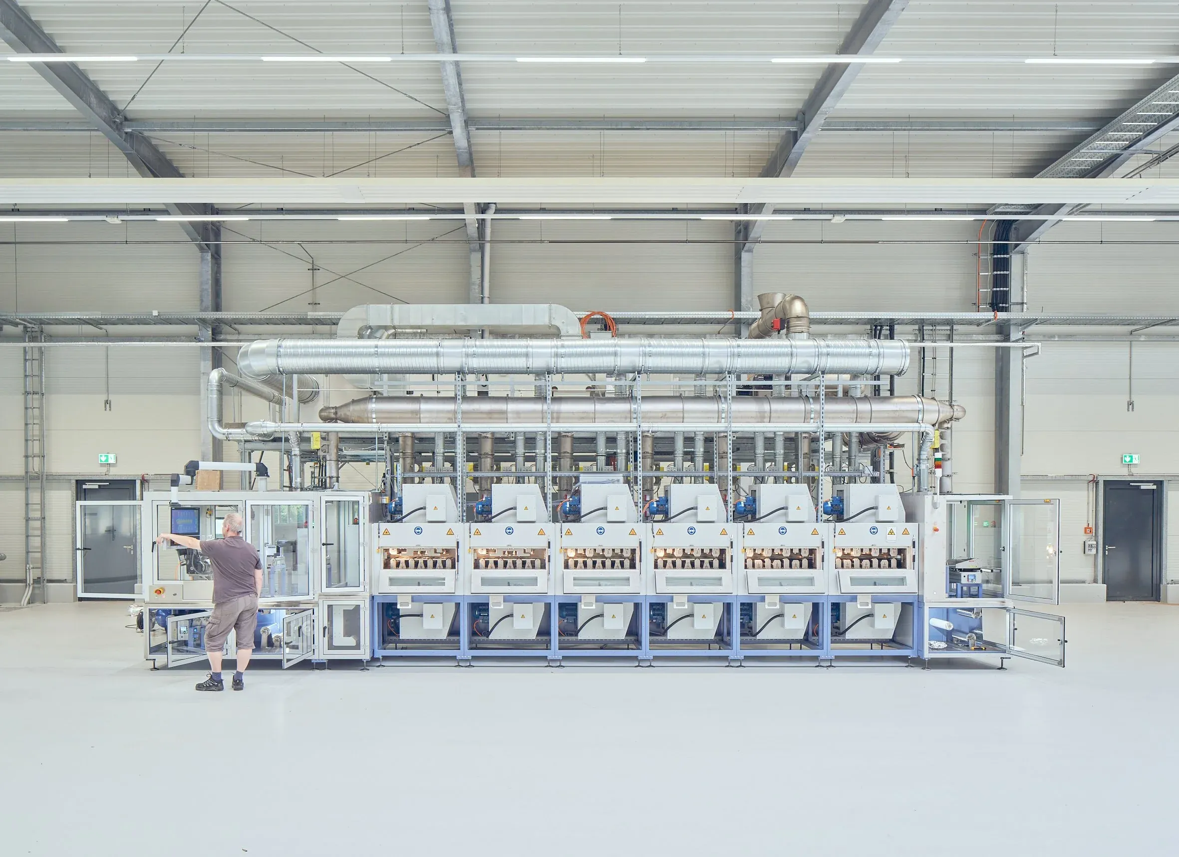Person bedient eine lange Produktionsmaschine mit Lüftungsrohren in der hellen Halle, Konstruktion im Hintergrund