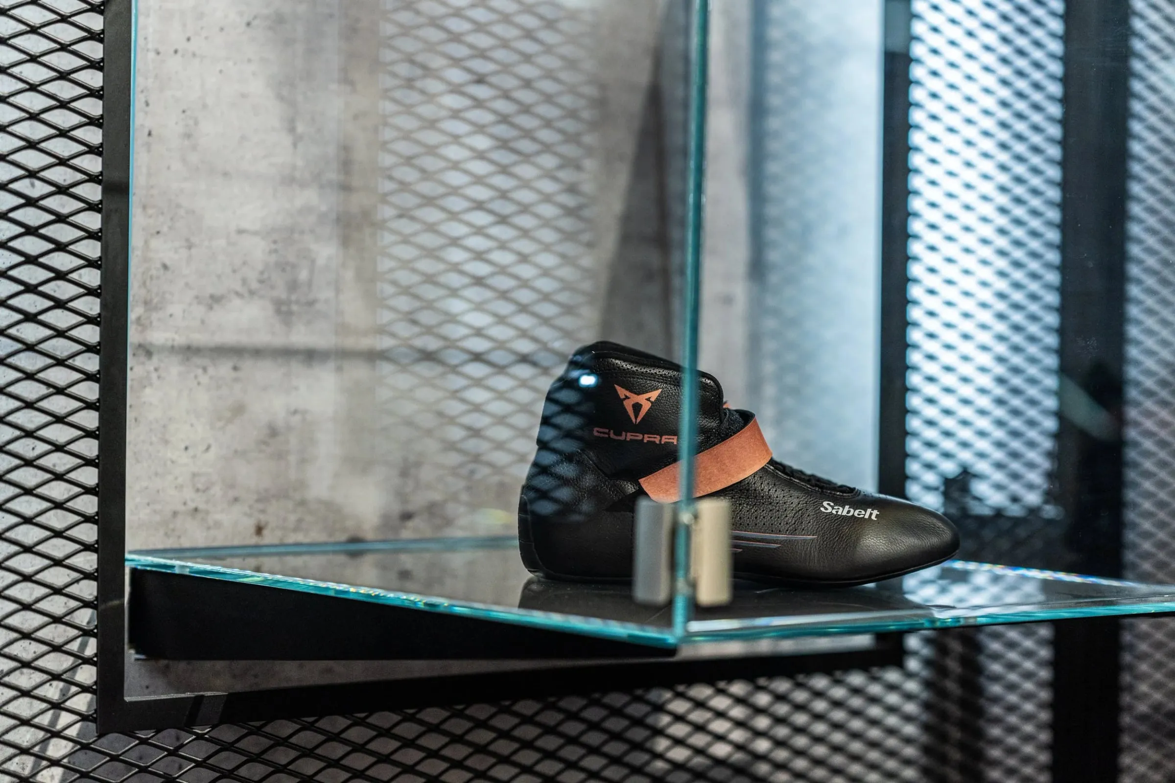 Schwarze Rennschuhe mit Cupra-Logo und kupfernem Band auf einer Glasplatte vor Sichtbetonwand