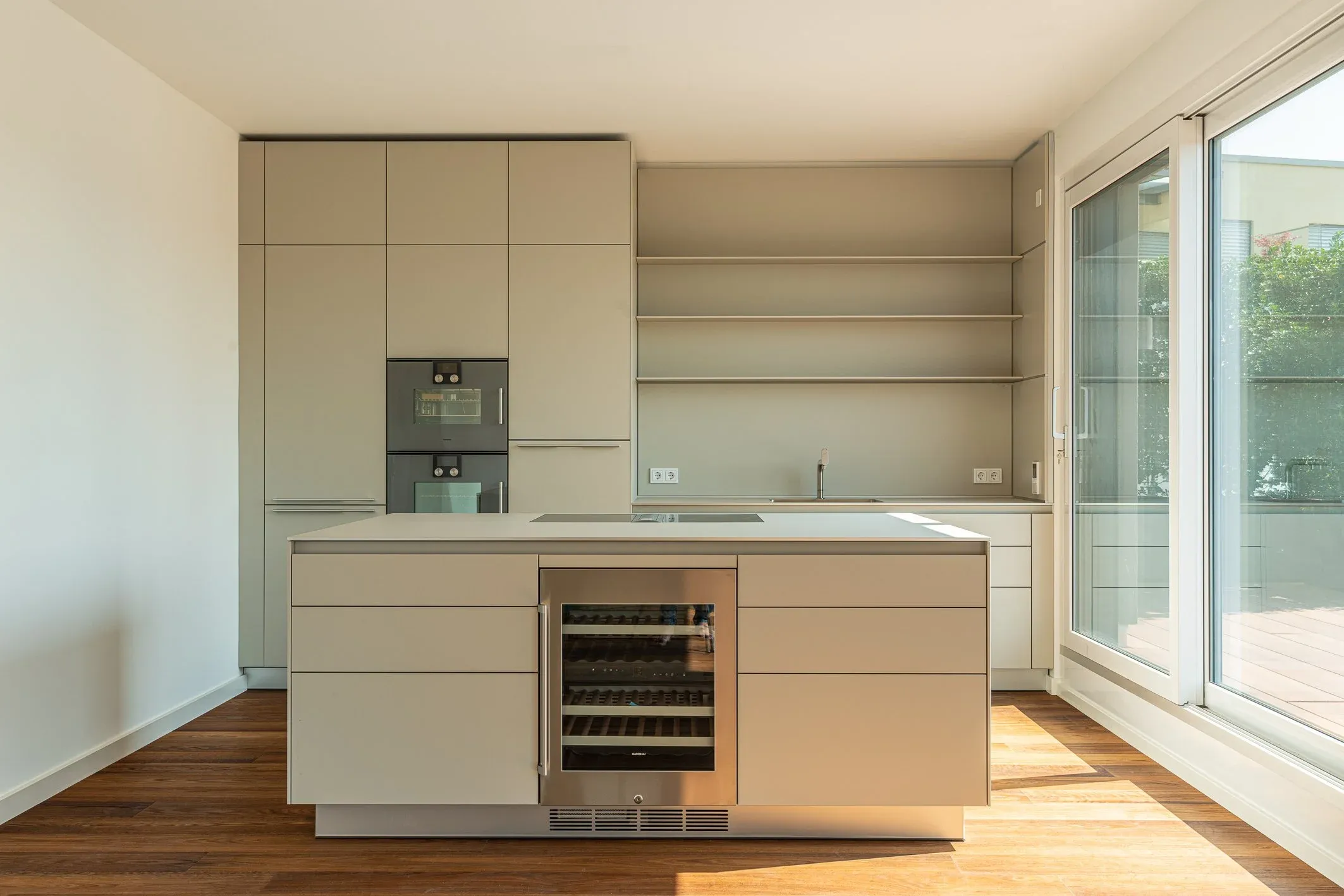 Innenaufnahme einer leeren Küche mit Kochinsel, Einbauschränken und bodentiefer Fensterfront zur Terrasse