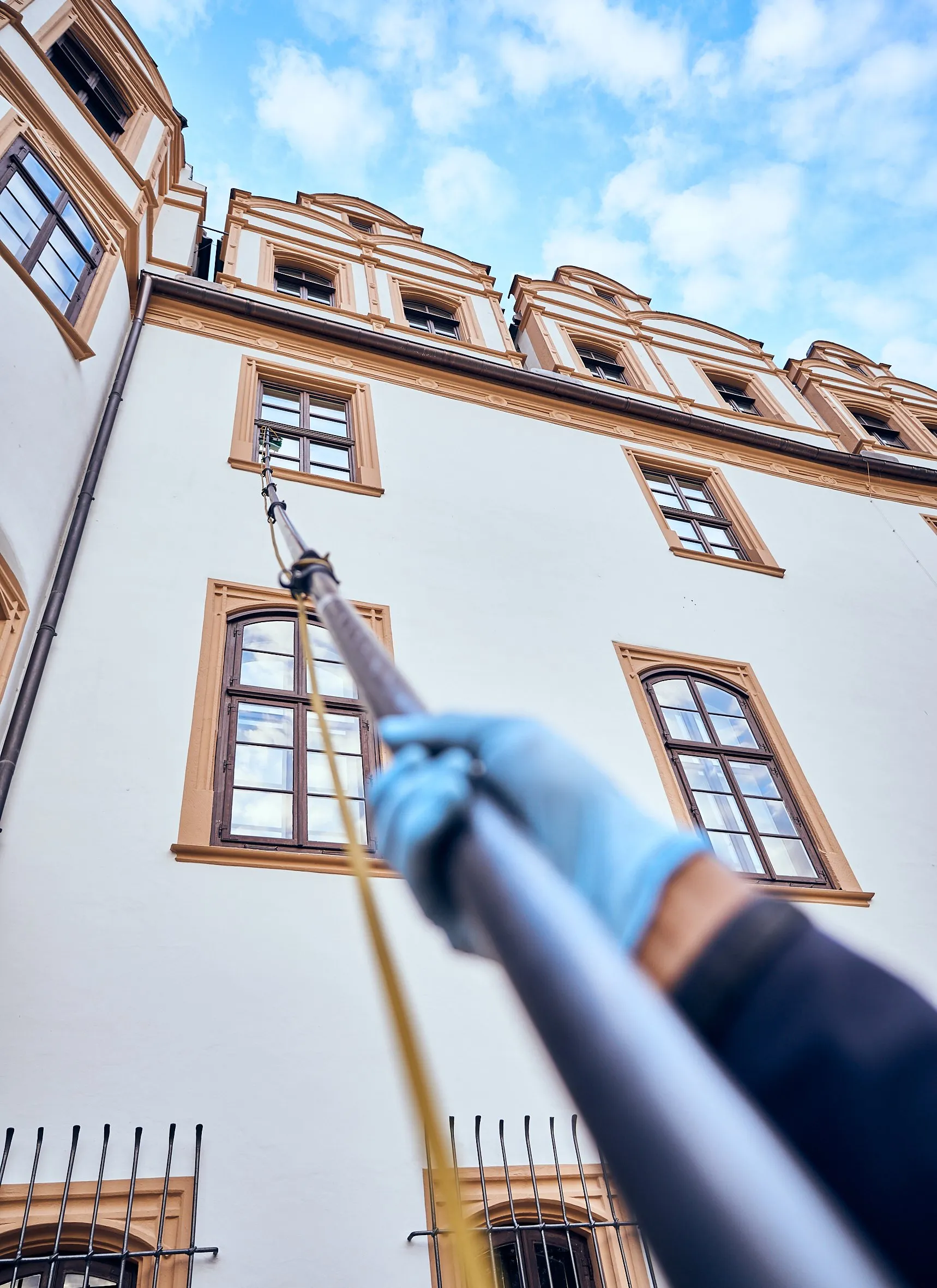 Untersicht, Mitarbeiter reinigt mit Teleskopstange ein hohes Fenster der Schlossfassade, blauer Himmel